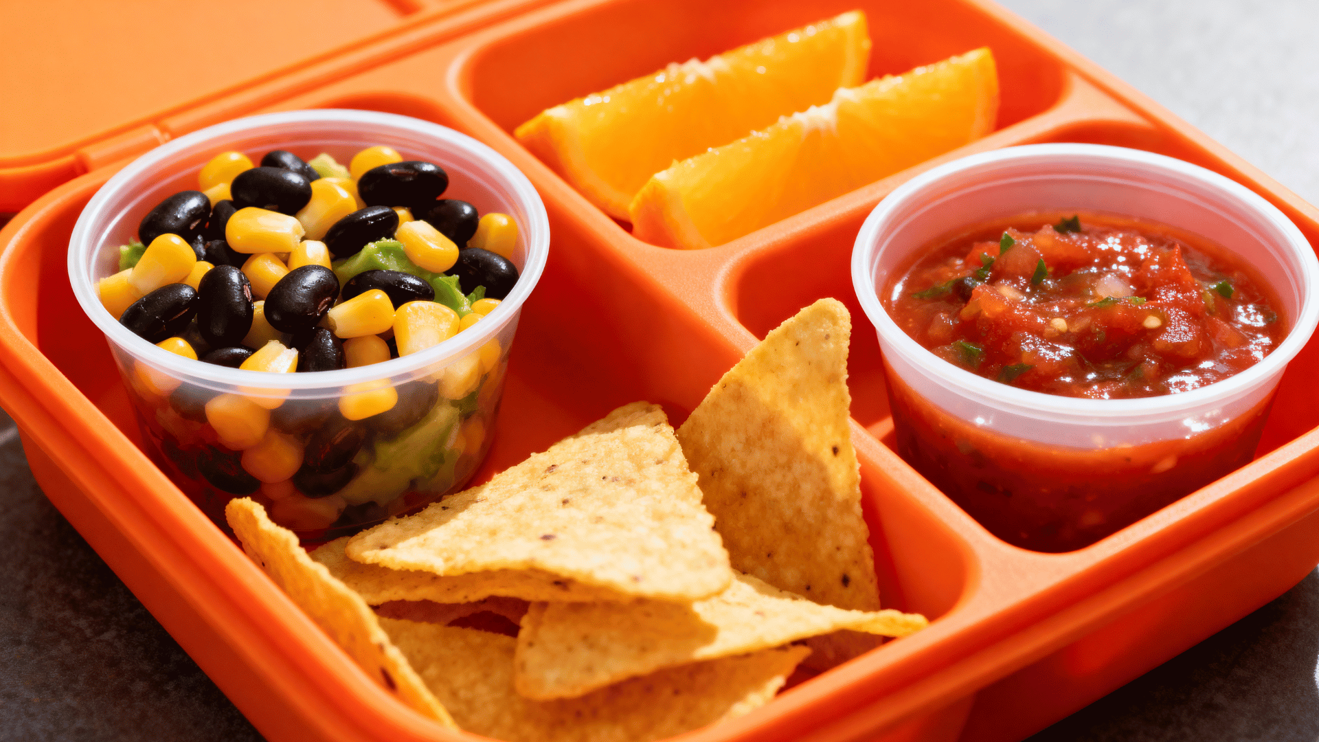 black bean and corn salad, tortilla chips, salsa, and orange wedges in a fiesta-orange lunchbox