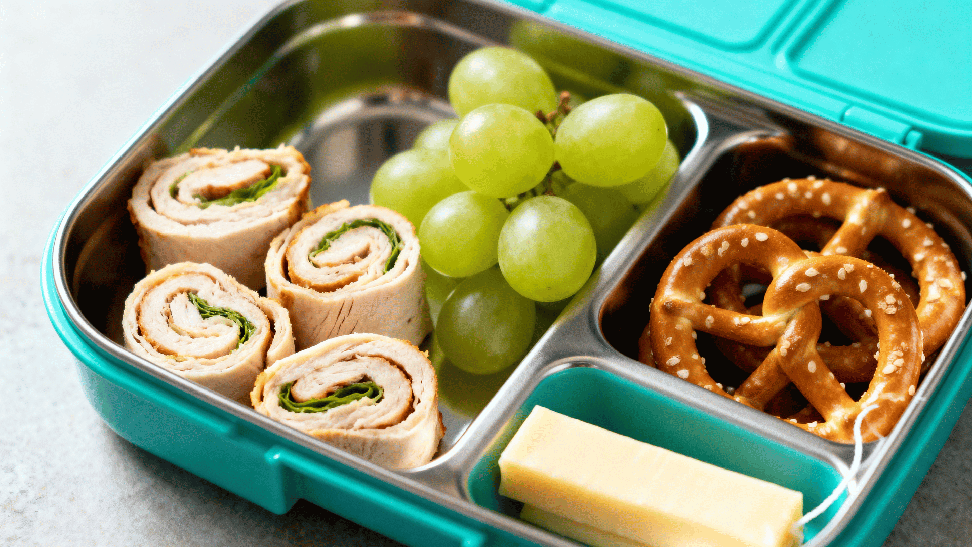 chicken roll-ups, grapes, pretzels, and string cheese in a turquoise divided lunchbox