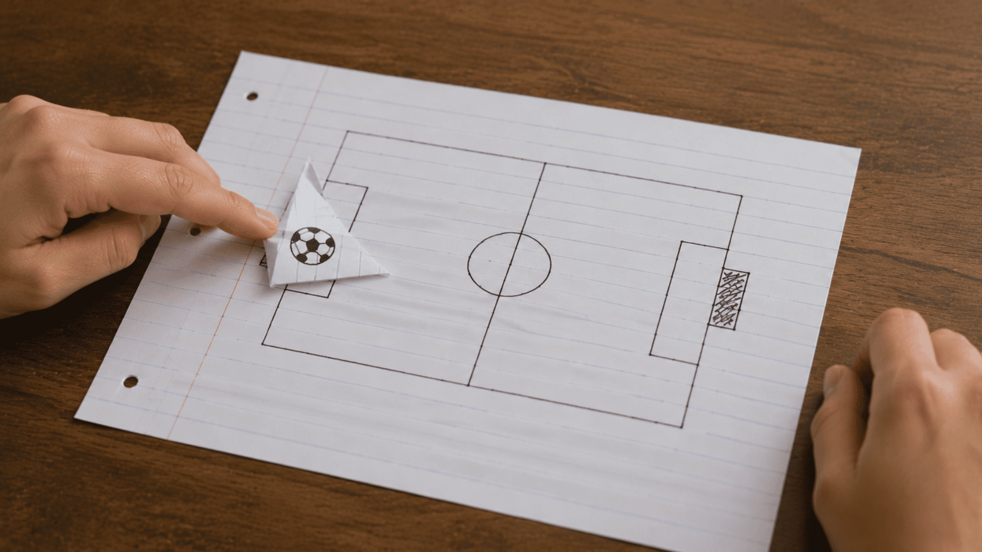 two hands playing a paper football game on a piece of lined paper with a hand-drawn soccer field outline