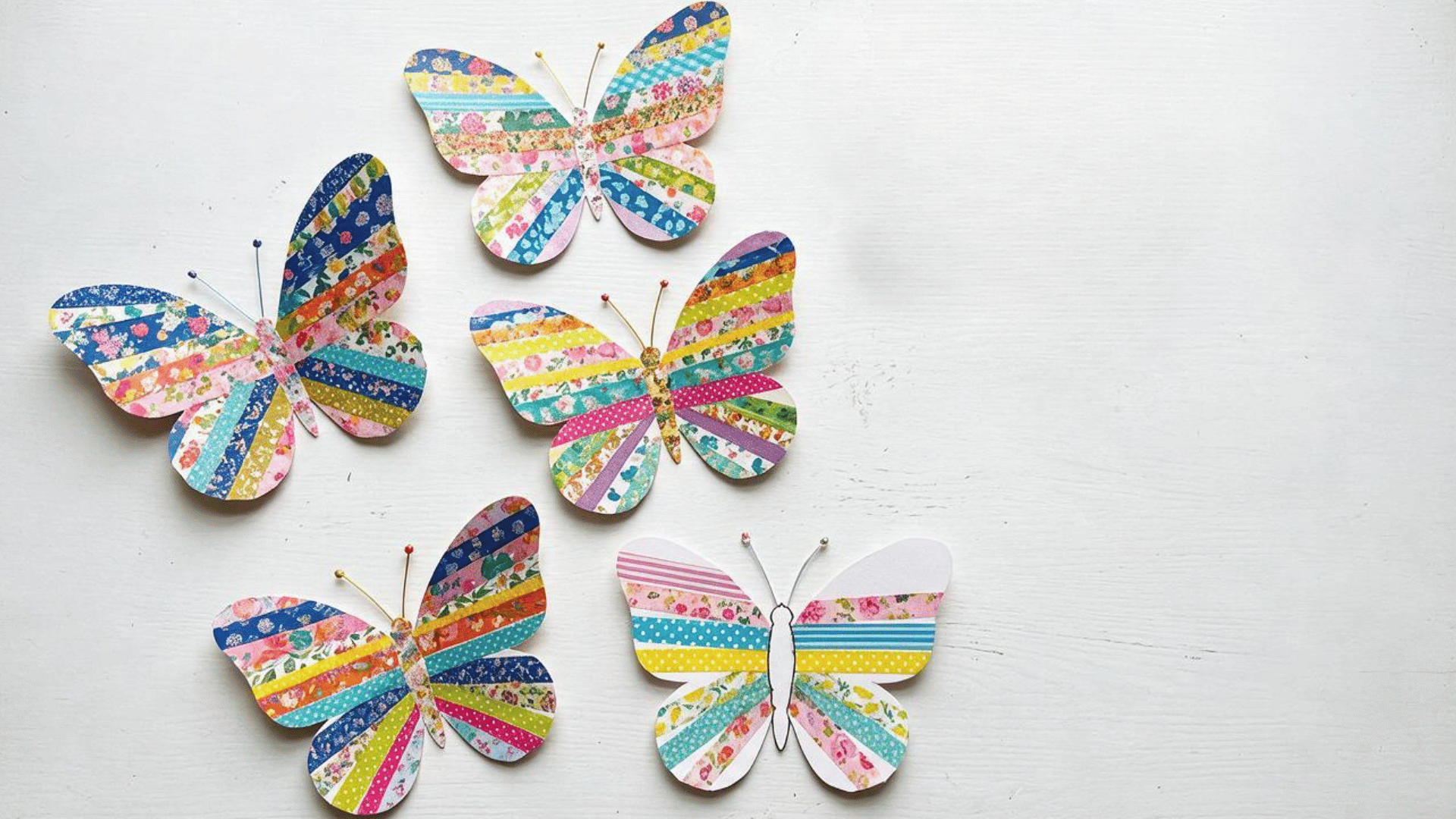 five colorful handmade paper butterflies with vibrant striped and floral patterns arranged on a white wooden surface, featuring bright pink, blue, yellow and green designs