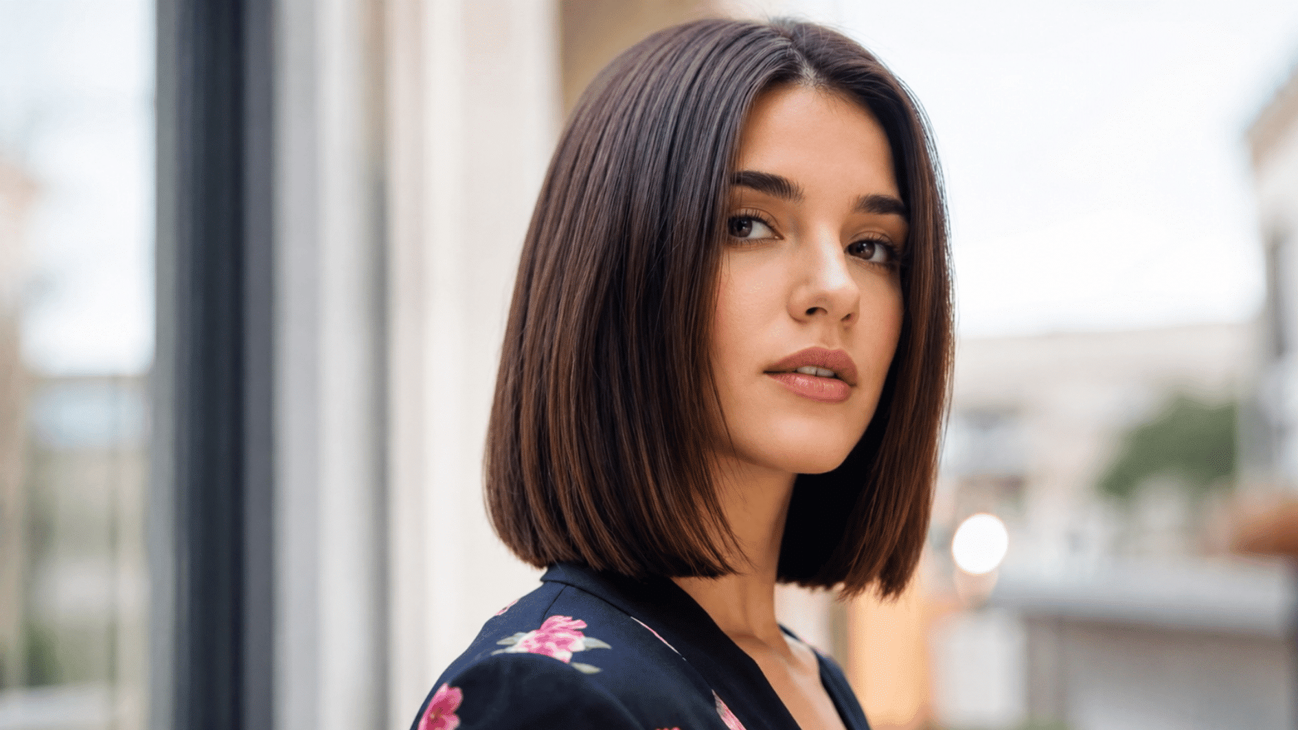 woman with sleek blunt bob haircut wearing a floral top near a bright outdoor street window