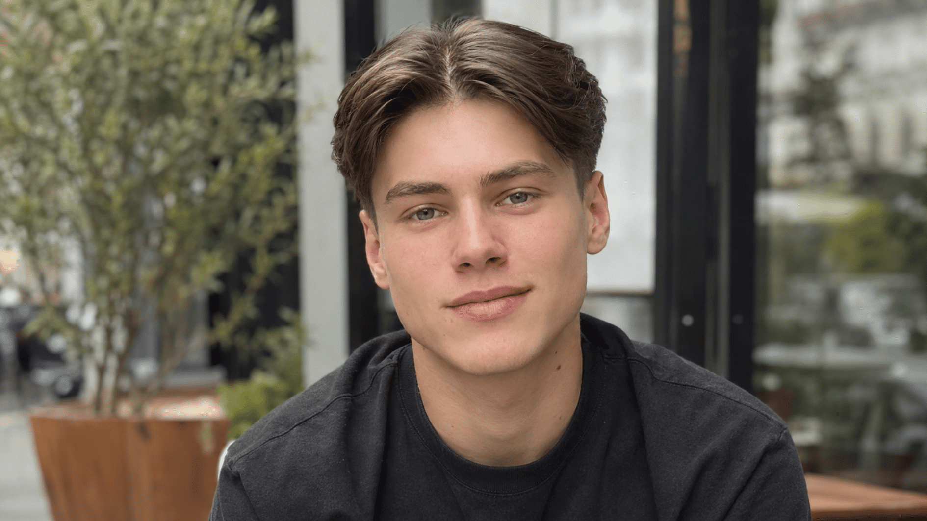 young man with middle part brown hair in a black shirt sitting near a glass window outside