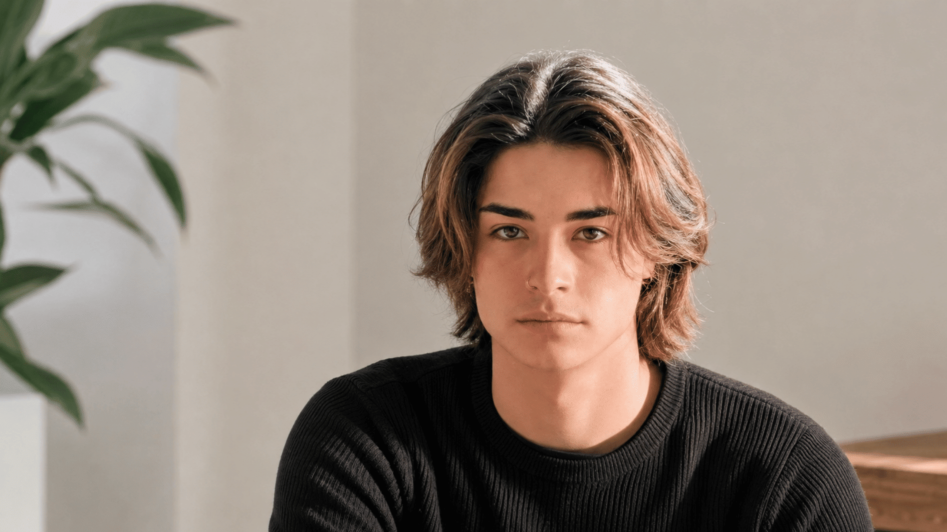 young man with shoulder-length middle part hair wearing a black sweater indoors near a plant