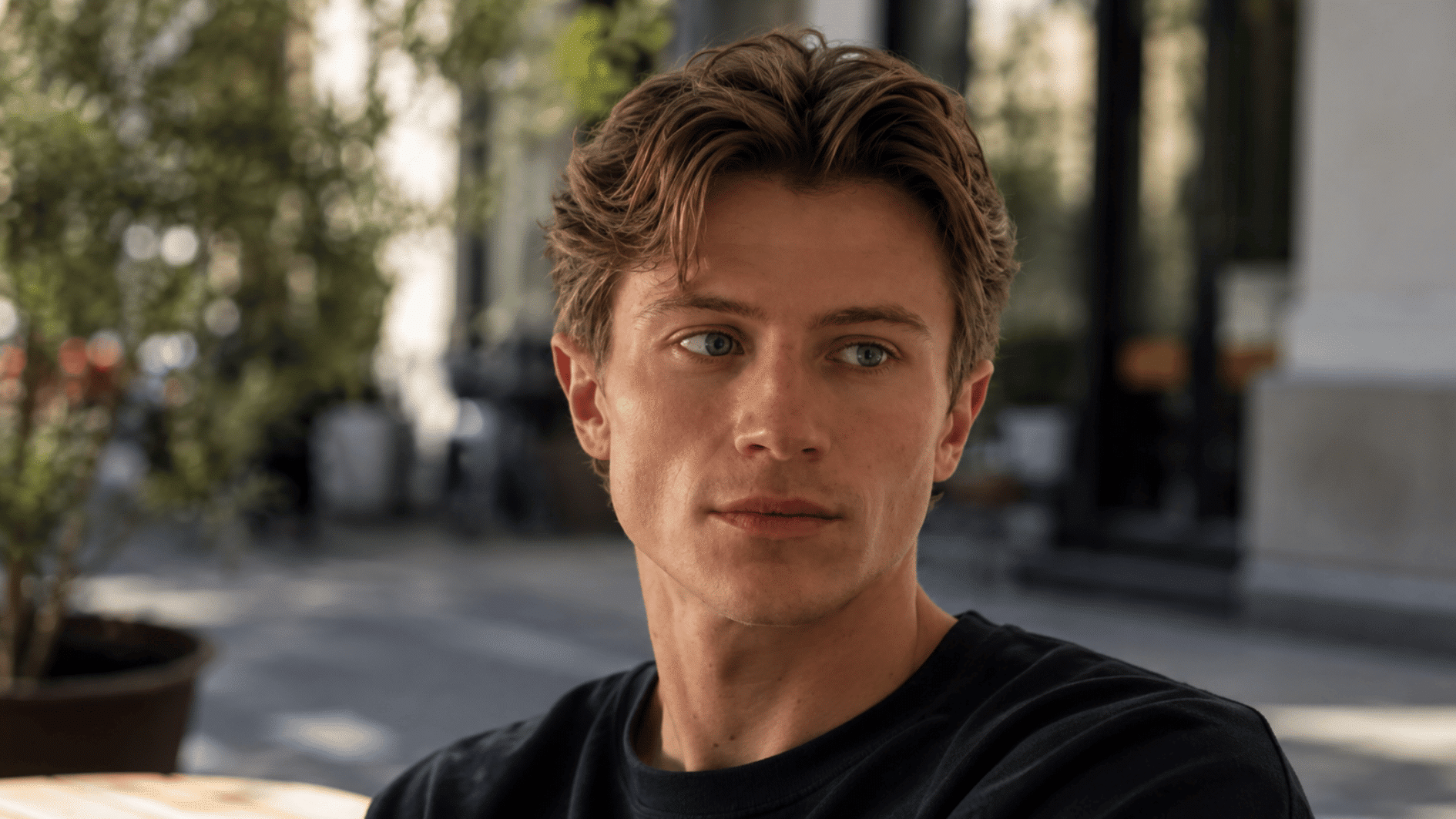 young man with swept brown middle part hair wearing a black shirt in an outdoor cafe setting