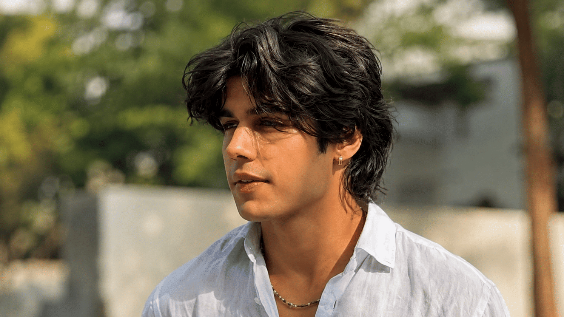 young man with thick wavy black hair wearing a white shirt in warm outdoor sunlight by greenery