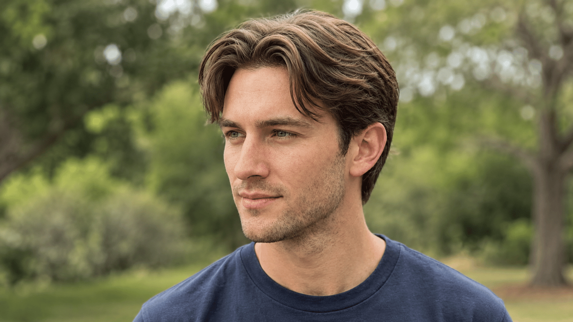 young man with wavy middle part hair wearing a navy shirt in a green outdoor park setting