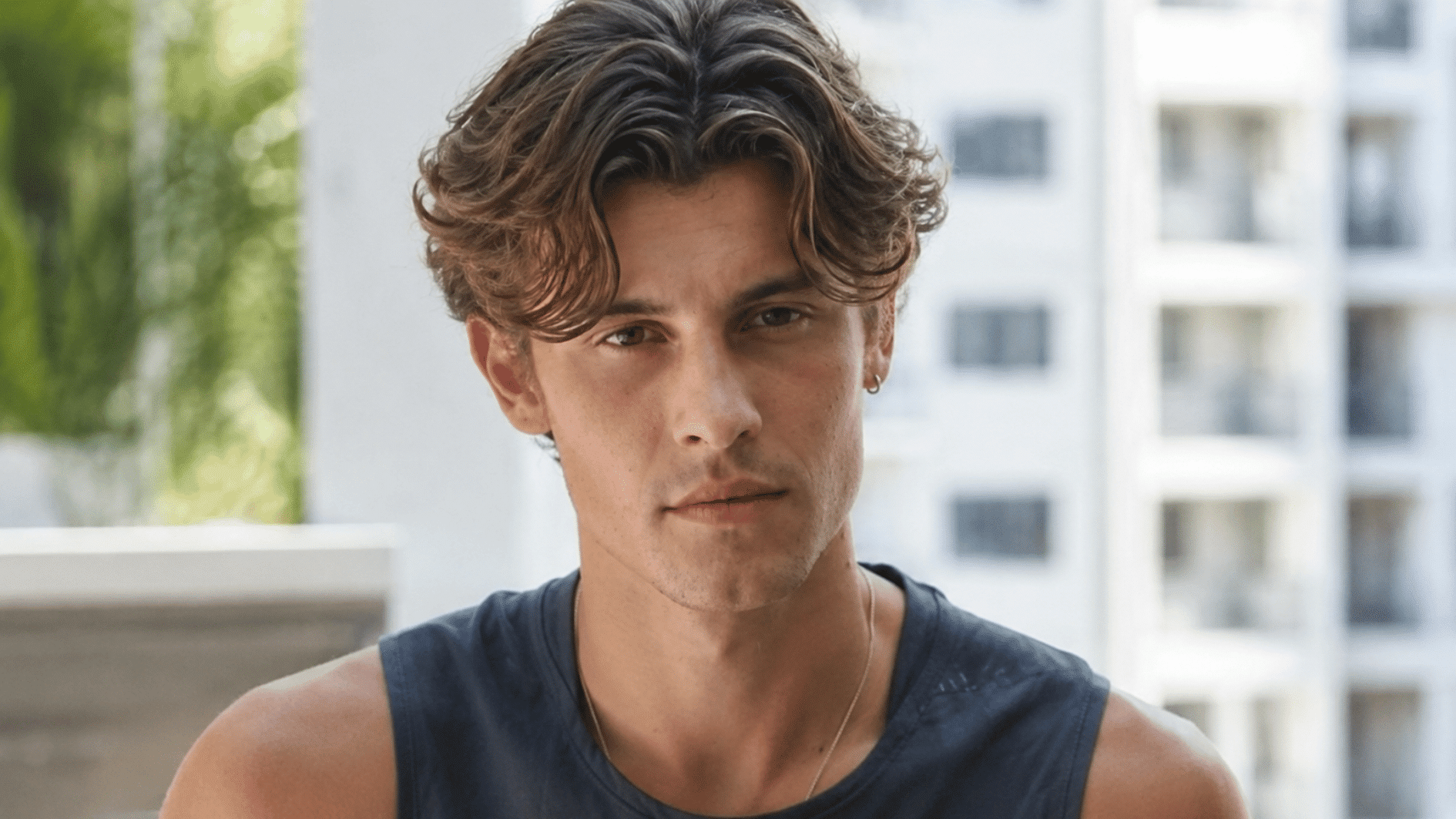 young man with wavy middle part hair wearing a sleeveless navy shirt near apartment windows