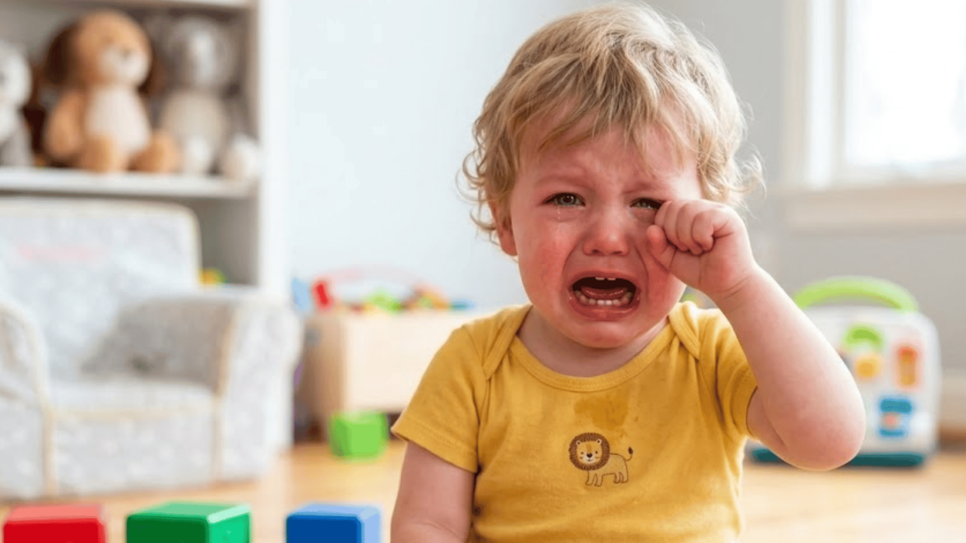 crying three-year-old child sitting on a playroom floor during a tantrum with colorful toy blocks nearby