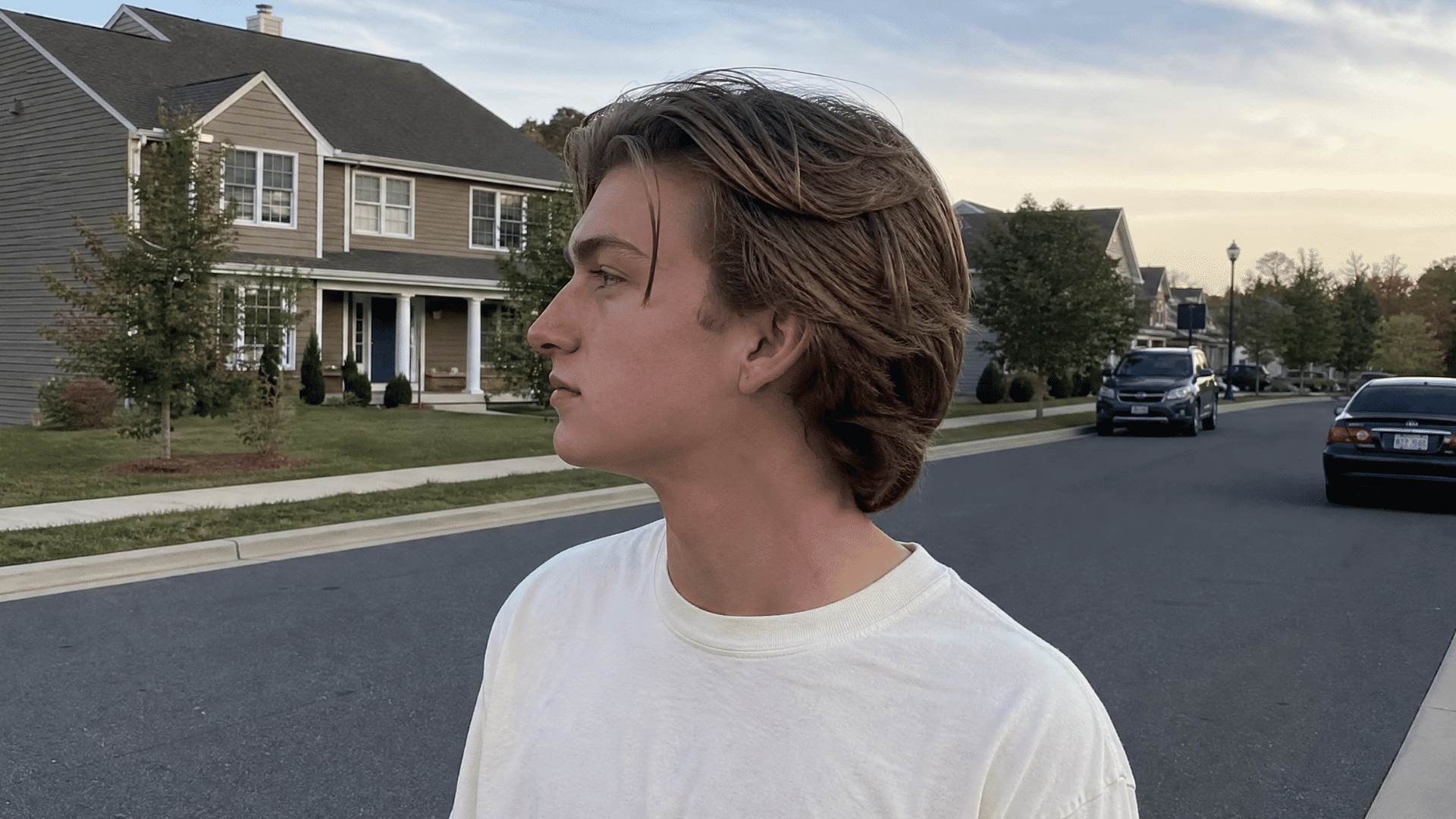 a boy with a bro flow, longer hair swept back off the face shown in side view on a suburban street