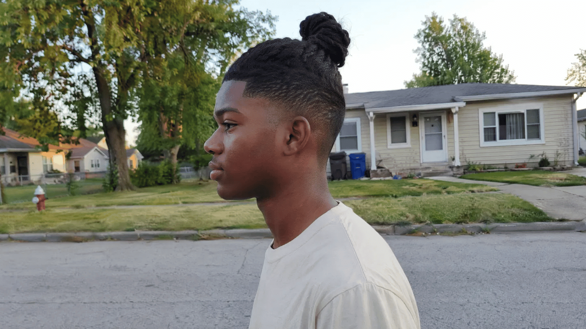 a boy with a top knot, hair tied up at the crown and faded sides shown in side view on a suburban street