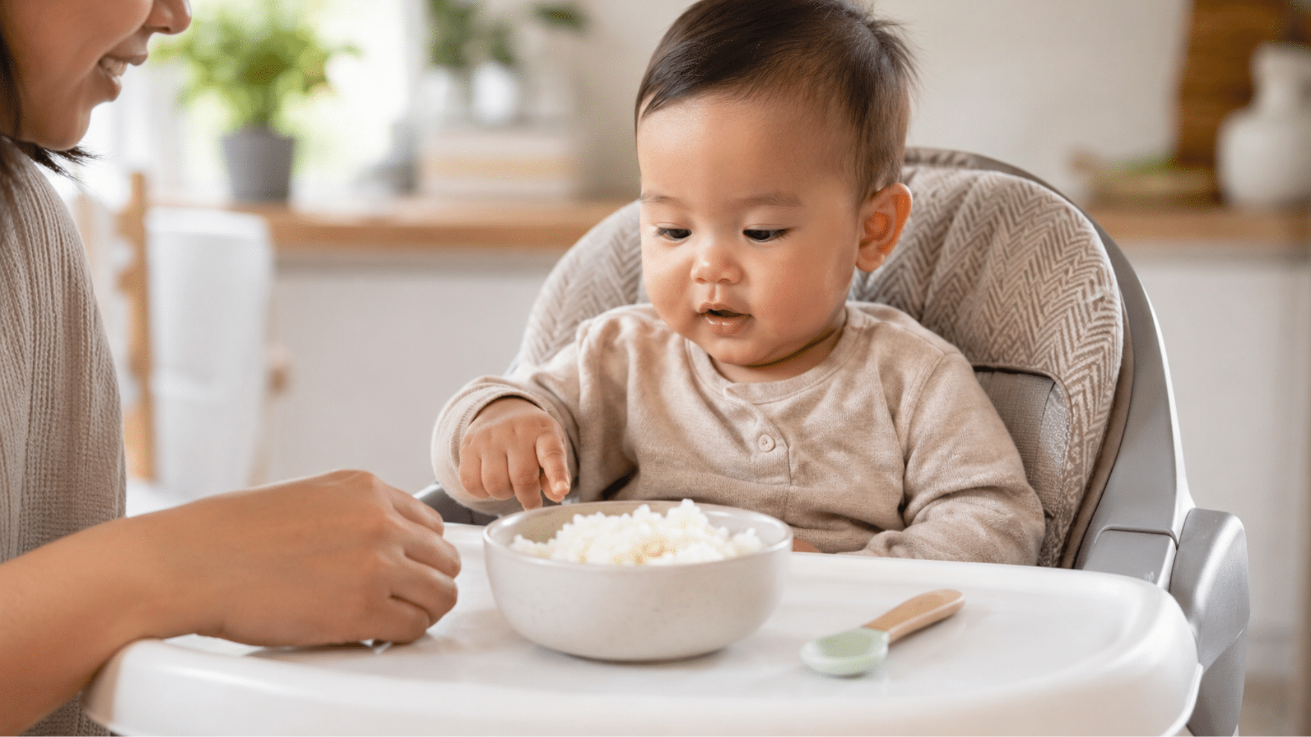 baby sitting in a high chair reaching toward a bowl of soft cooked rice as a parent watches nearby