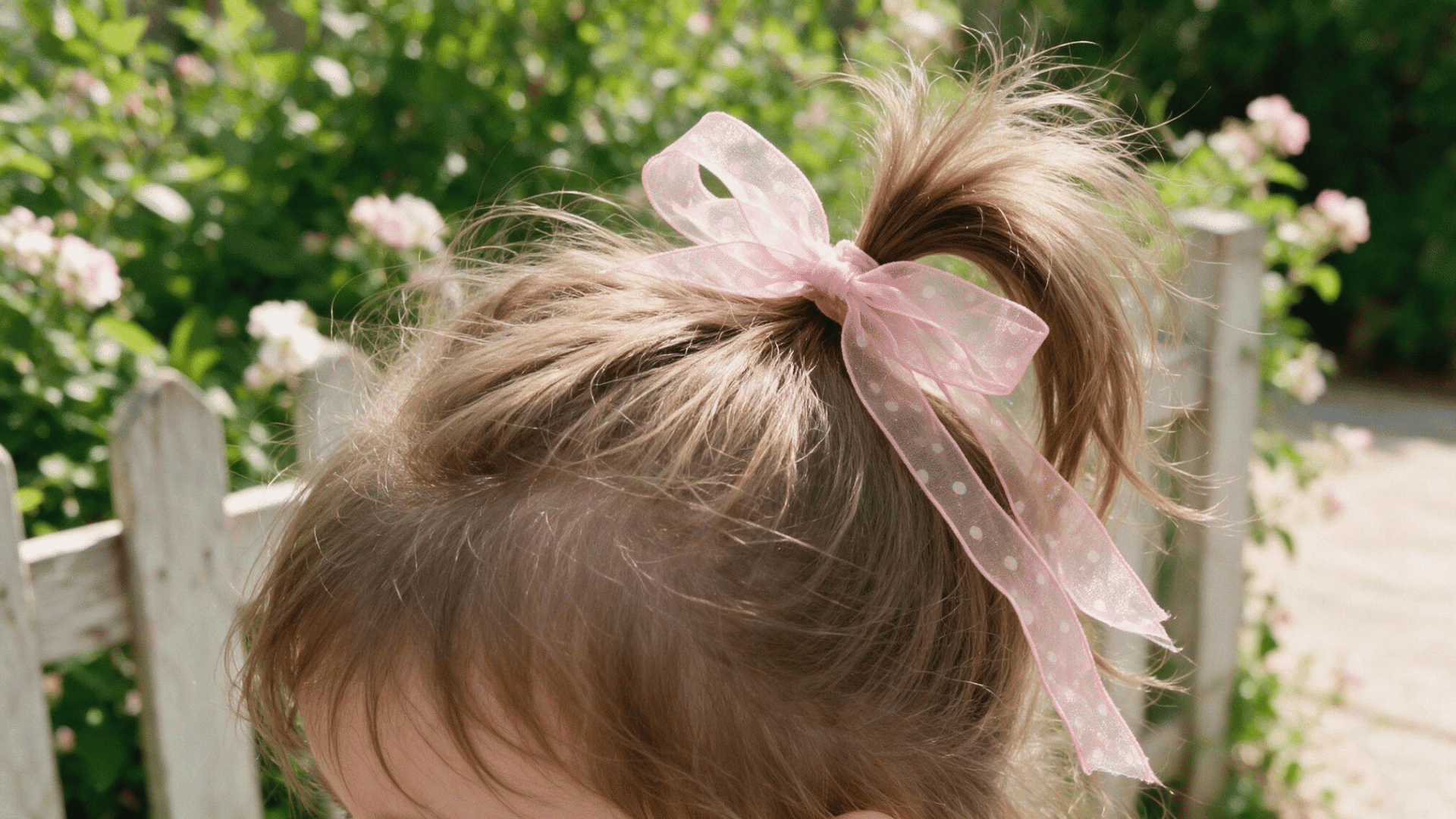 baby hairstyle with a high ponytail finished with a soft pink ribbon bow in natural outdoor sunlight beside a blooming garden fence
