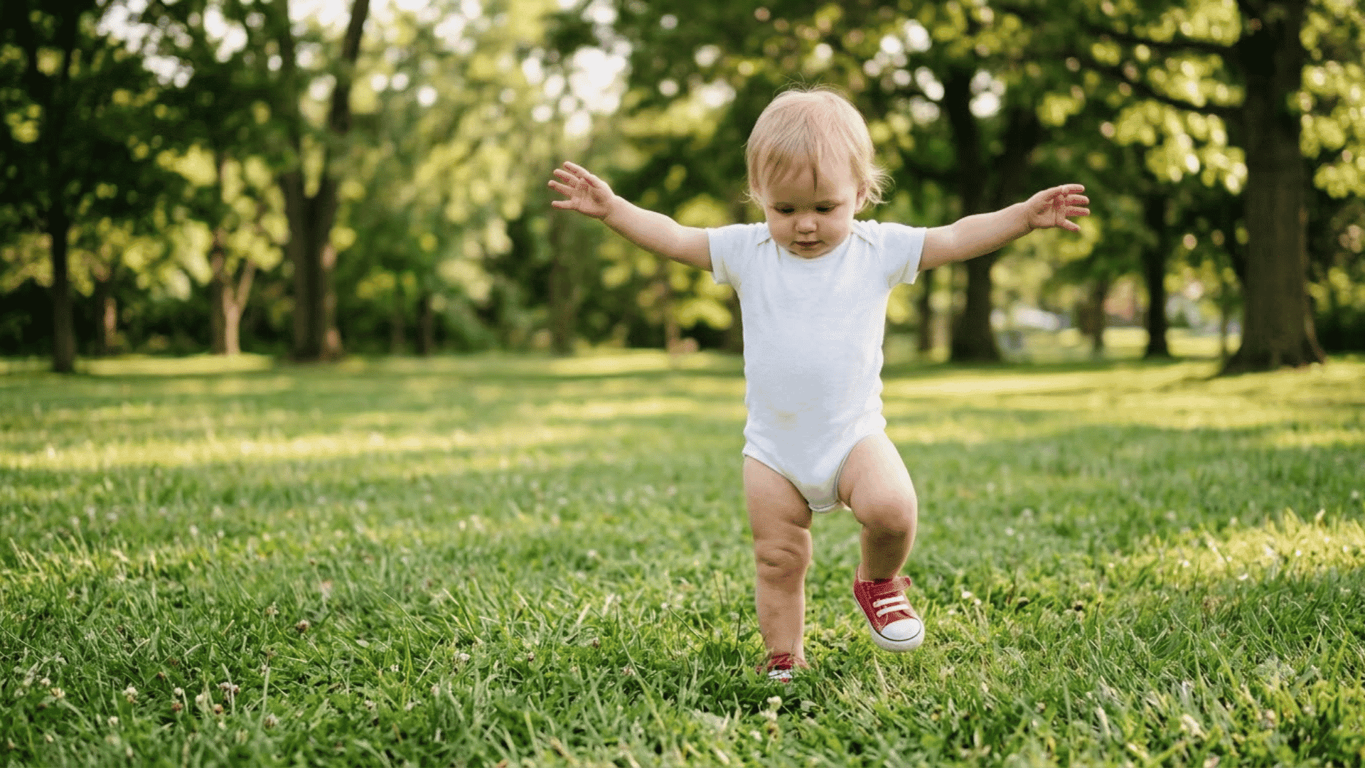 baby taking unsteady first steps alone on a sunny lawn with both arms extended outward for balance