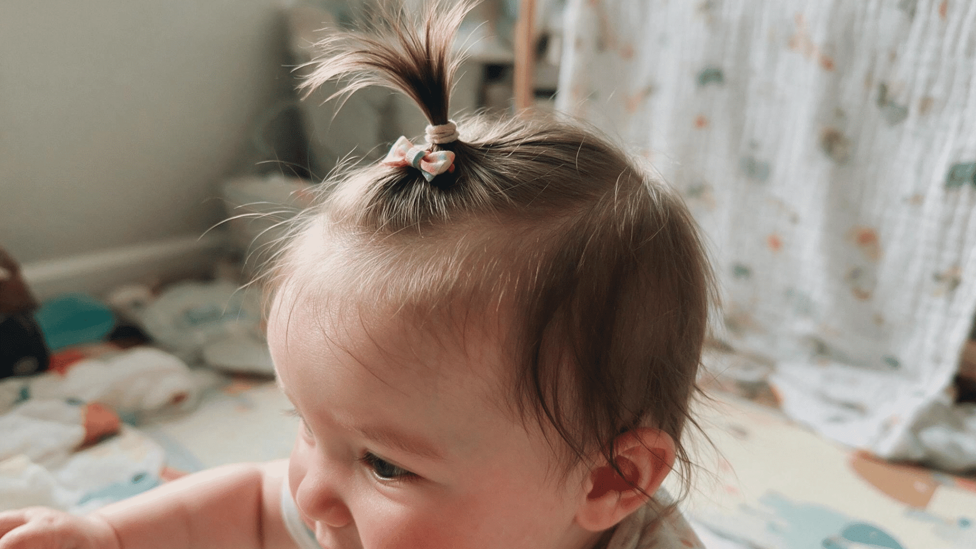 baby with a tiny top ponytail secured by a pastel bow sitting indoors with soft natural light and a cozy nursery background