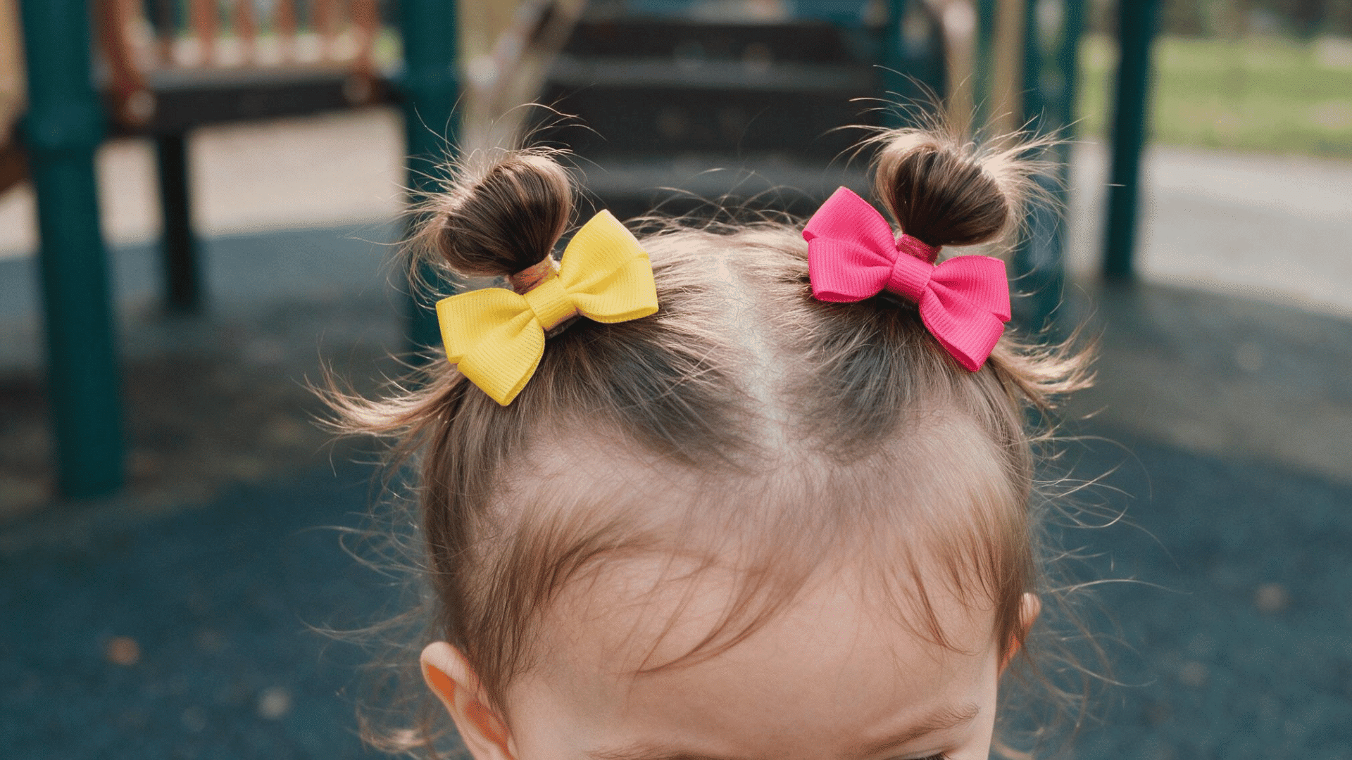 baby with double mini buns decorated with colorful bows at an outdoor playground in soft natural daylight viewed from the front