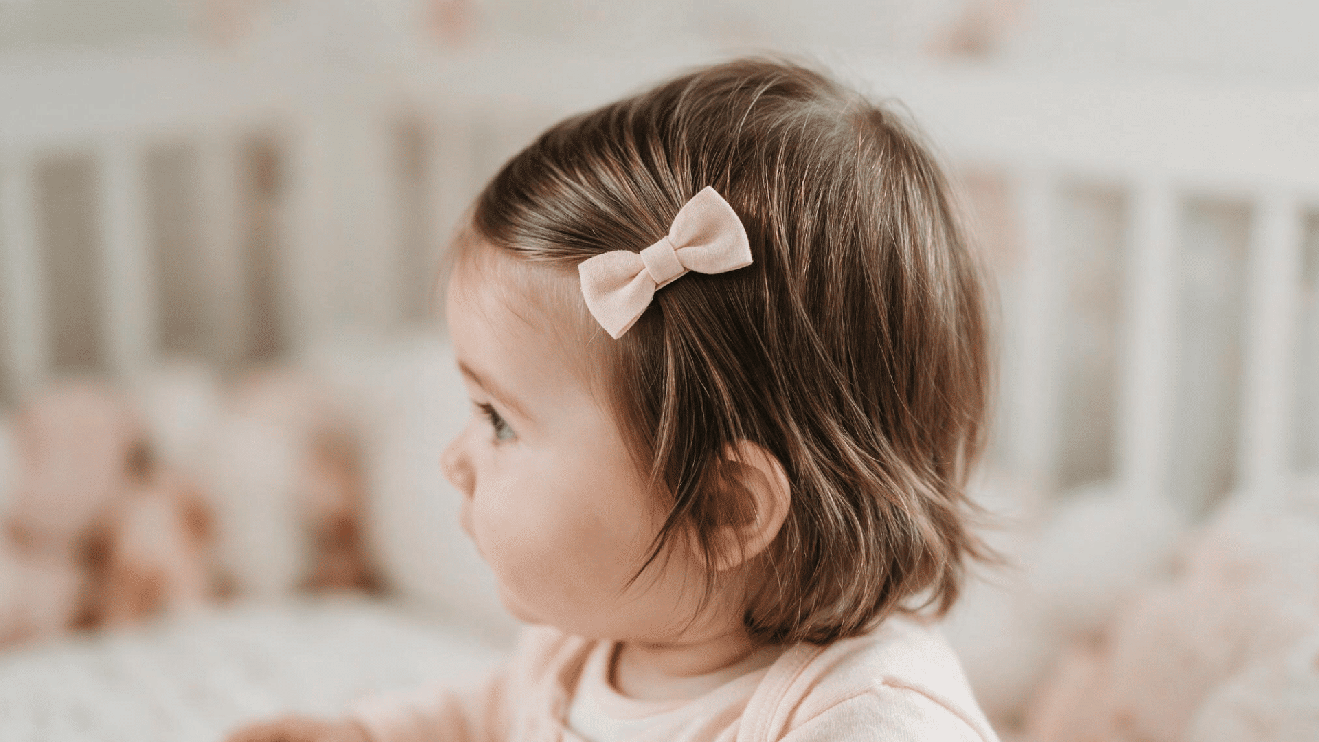 baby with soft brown hair swept to the side and styled with a small beige bow clip while sitting in a cozy nursery