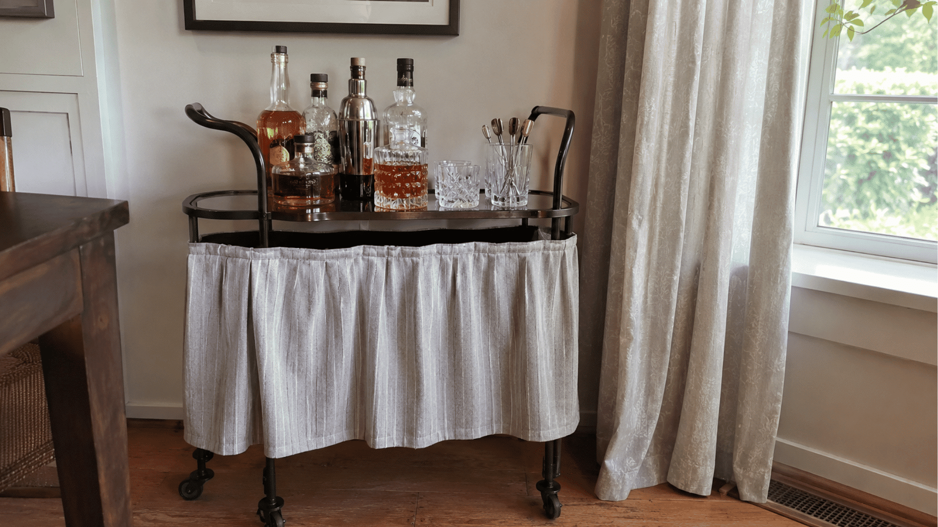 Bar cart with a pleated gray fabric skirt hiding the base, topped with whiskey bottles, shaker, and glasses