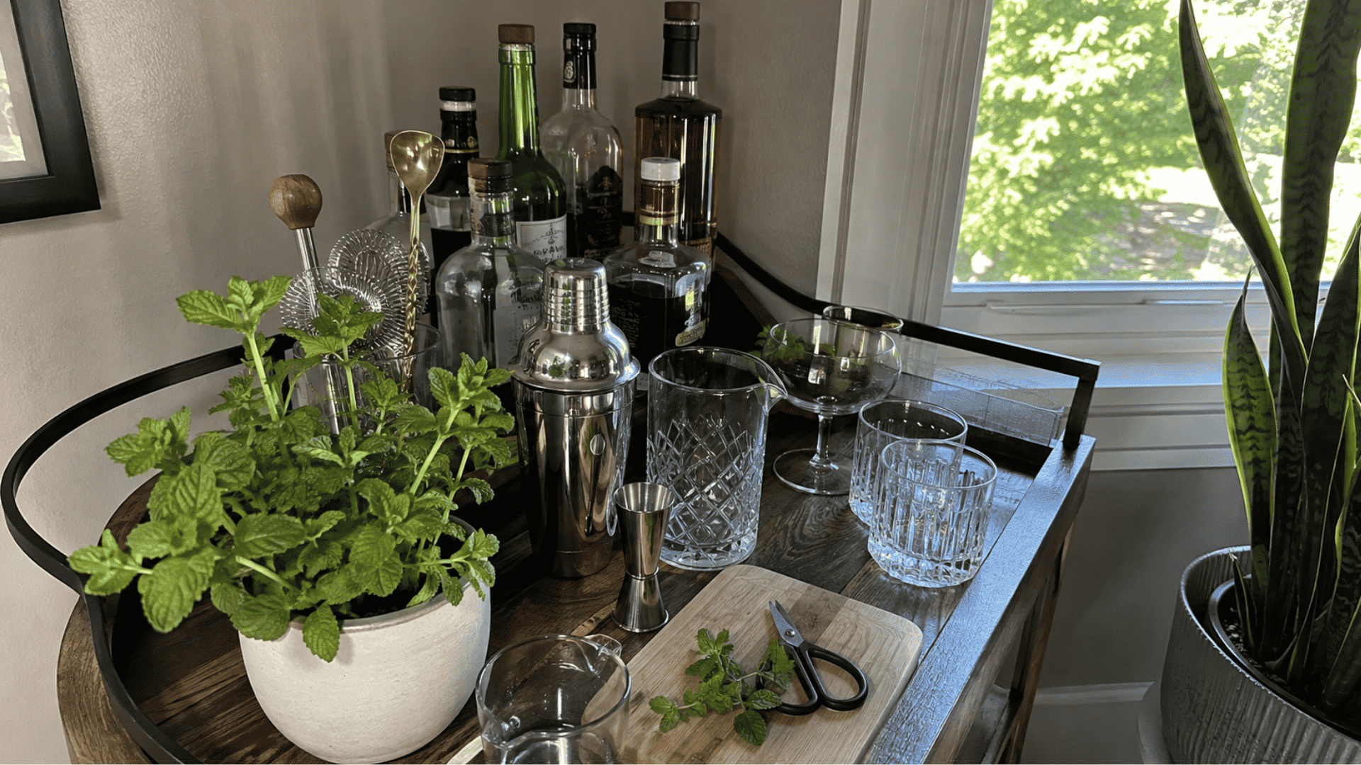 Bar cart with a potted mint plant, cocktail shaker, jigger, bottles, mixing glass, and fresh mint on cutting board