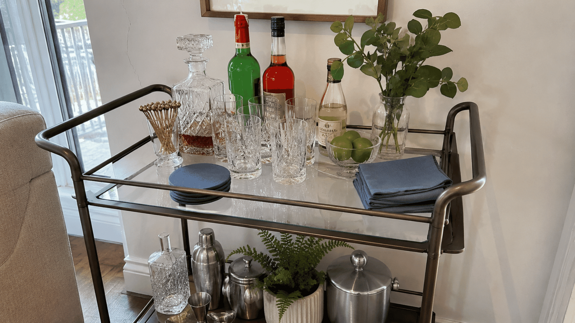 Bar cart with blue coasters and napkins, decanter, crystal glasses, limes, greenery, and colorful liquor bottles