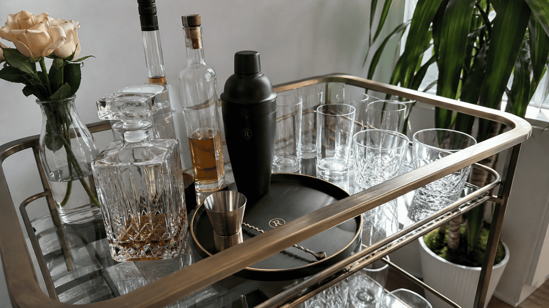 Bar cart with monogrammed shaker and tray, decanter, whiskey bottles, glasses, roses, and a small potted plant