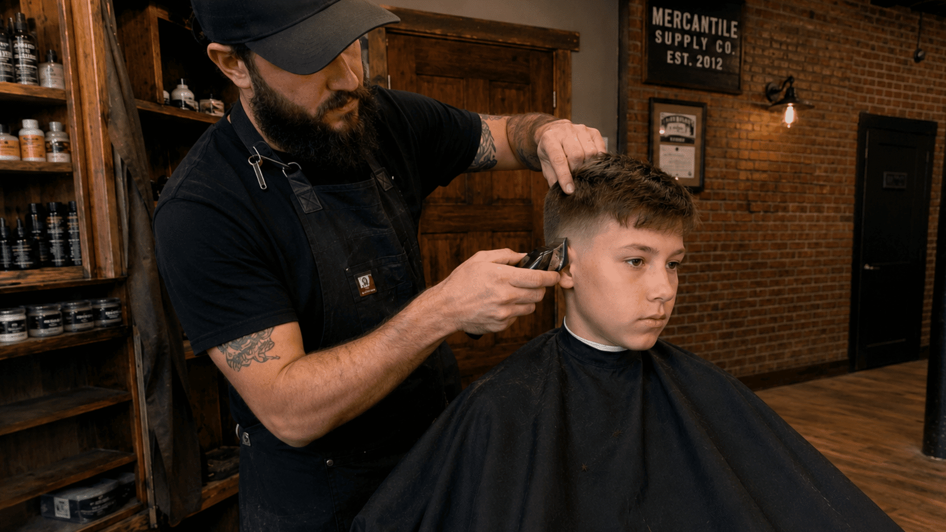 boy getting boys crew cut haircut from barber with clippers inside rustic mercantile supply barbershop