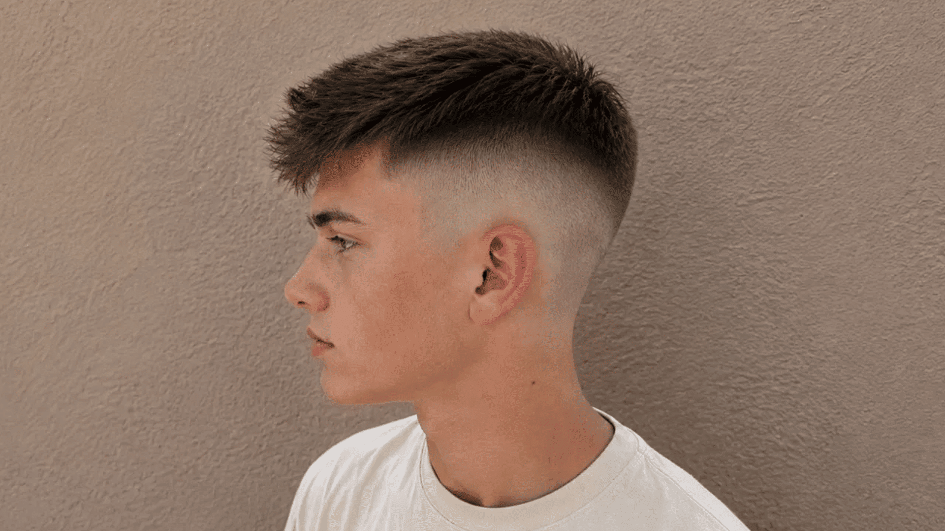 boy in profile with a high fade haircut, wearing a white t-shirt against a beige wall