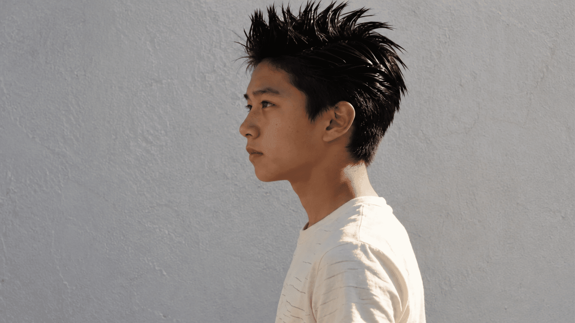 boy in profile with a spiky hairstyle, wearing a cream t-shirt against a grey wall