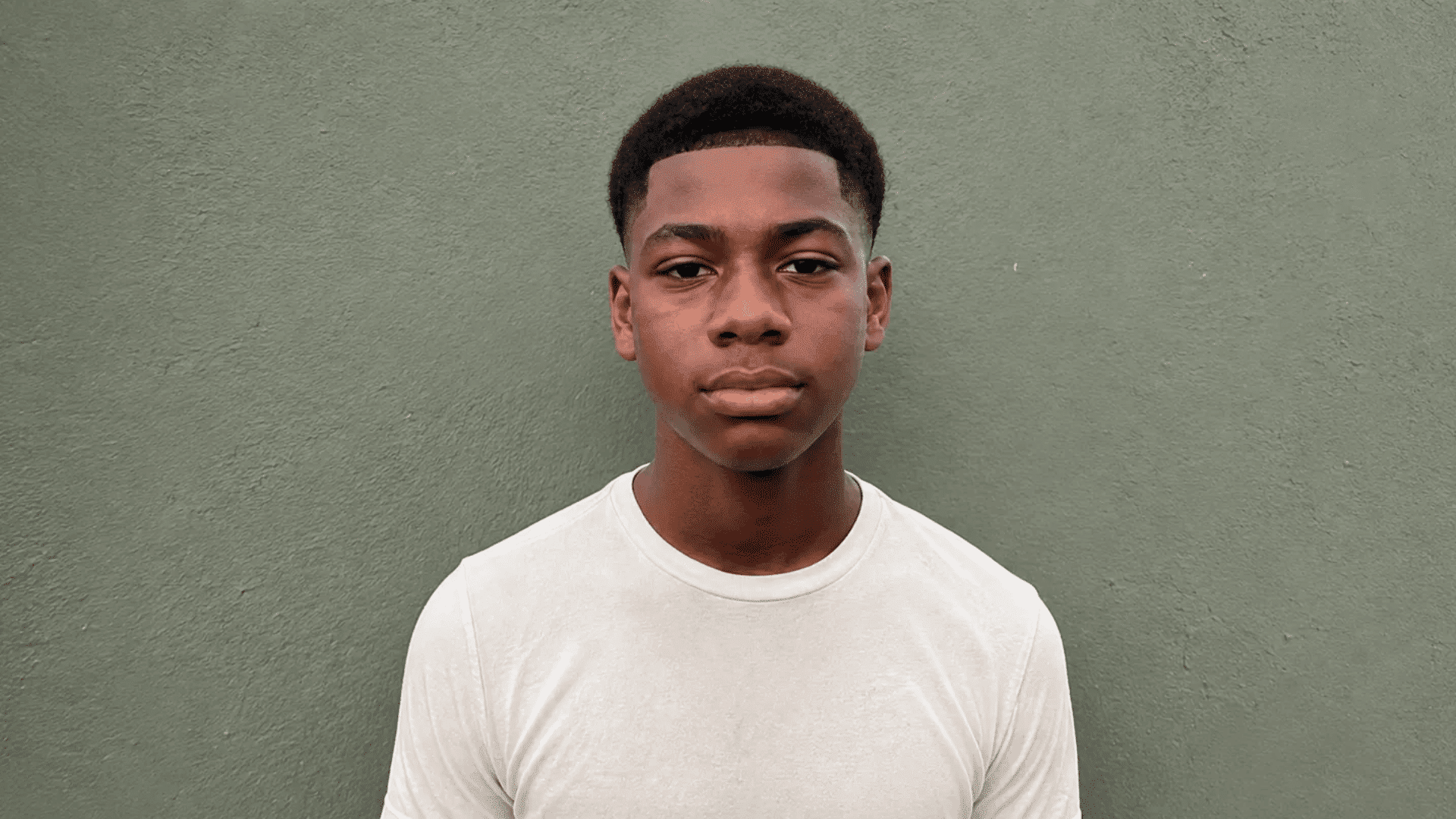 boy with a line up and short top haircut, wearing a white t-shirt against a sage green wall
