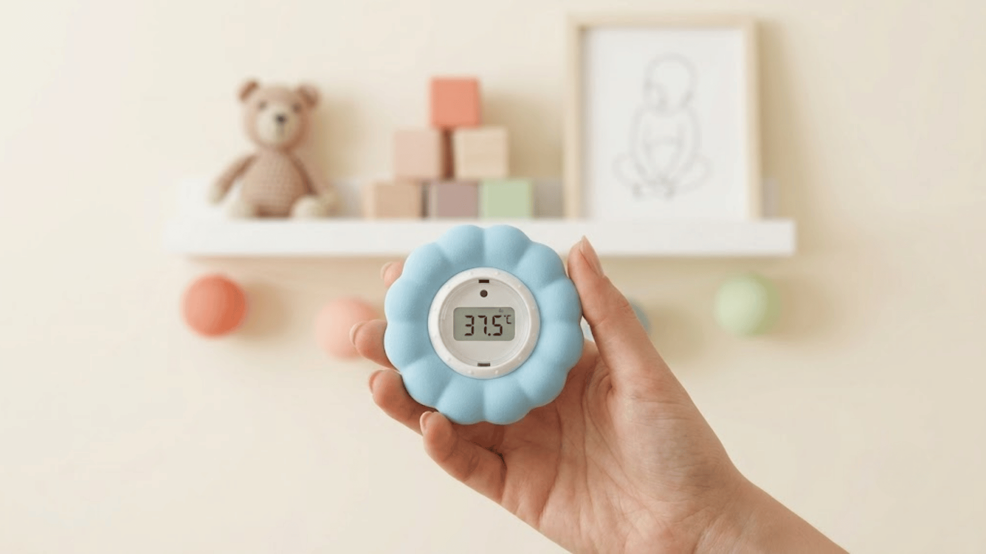 hand holds a digital nursery thermometer showing 37.5°C against a softly decorated baby room wall indoors