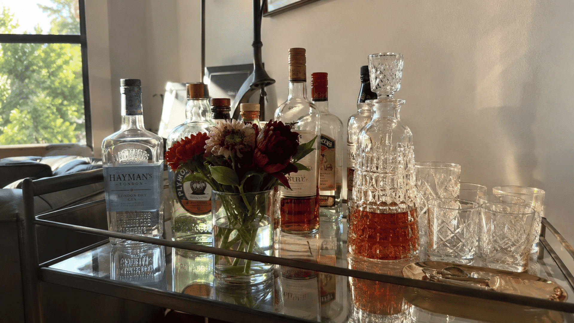 Glass bar cart with gin, whiskey bottles, crystal decanter, rocks glasses, and a small vase of red and white flowers