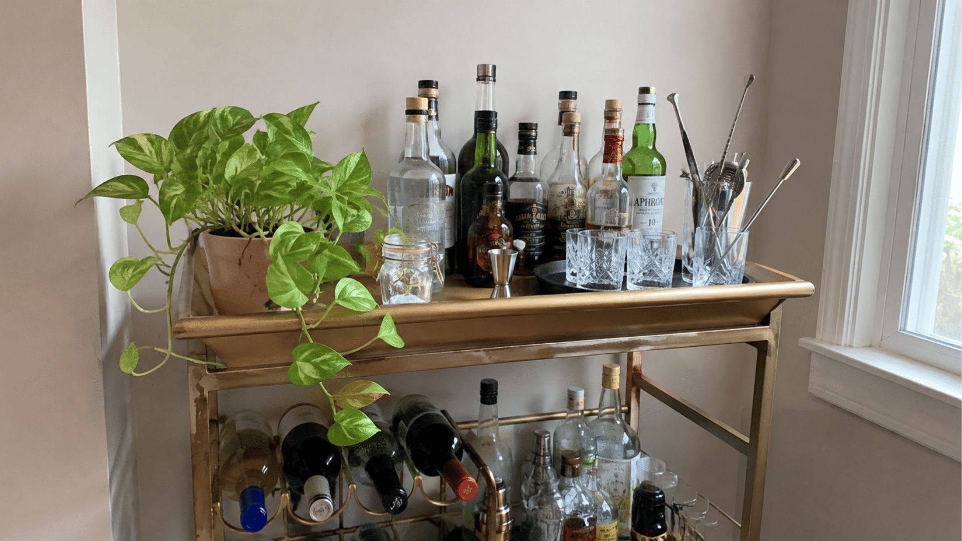 Gold bar cart with a trailing pothos plant, whiskey bottles, jigger, glasses, and a wine bottle rack below