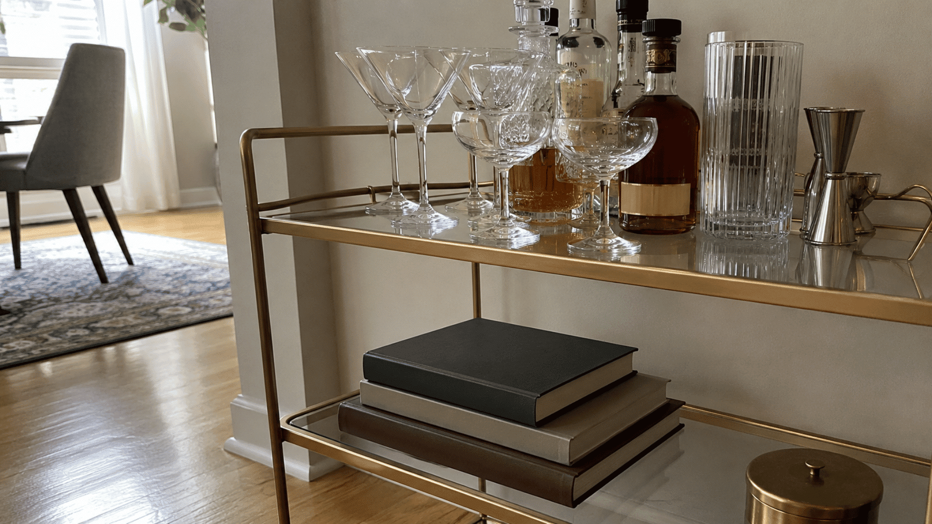 Gold bar cart with martini and coupe glasses, decanters, jigger, and a stack of hardcover books on lower shelf