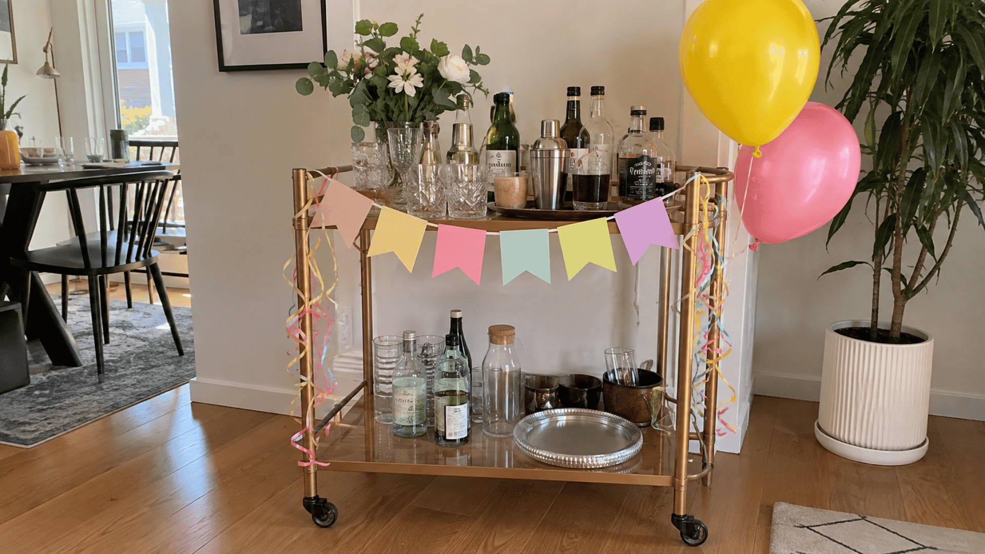 Gold rolling bar cart decorated with a pastel banner, streamers, balloons, flowers, and party bottles