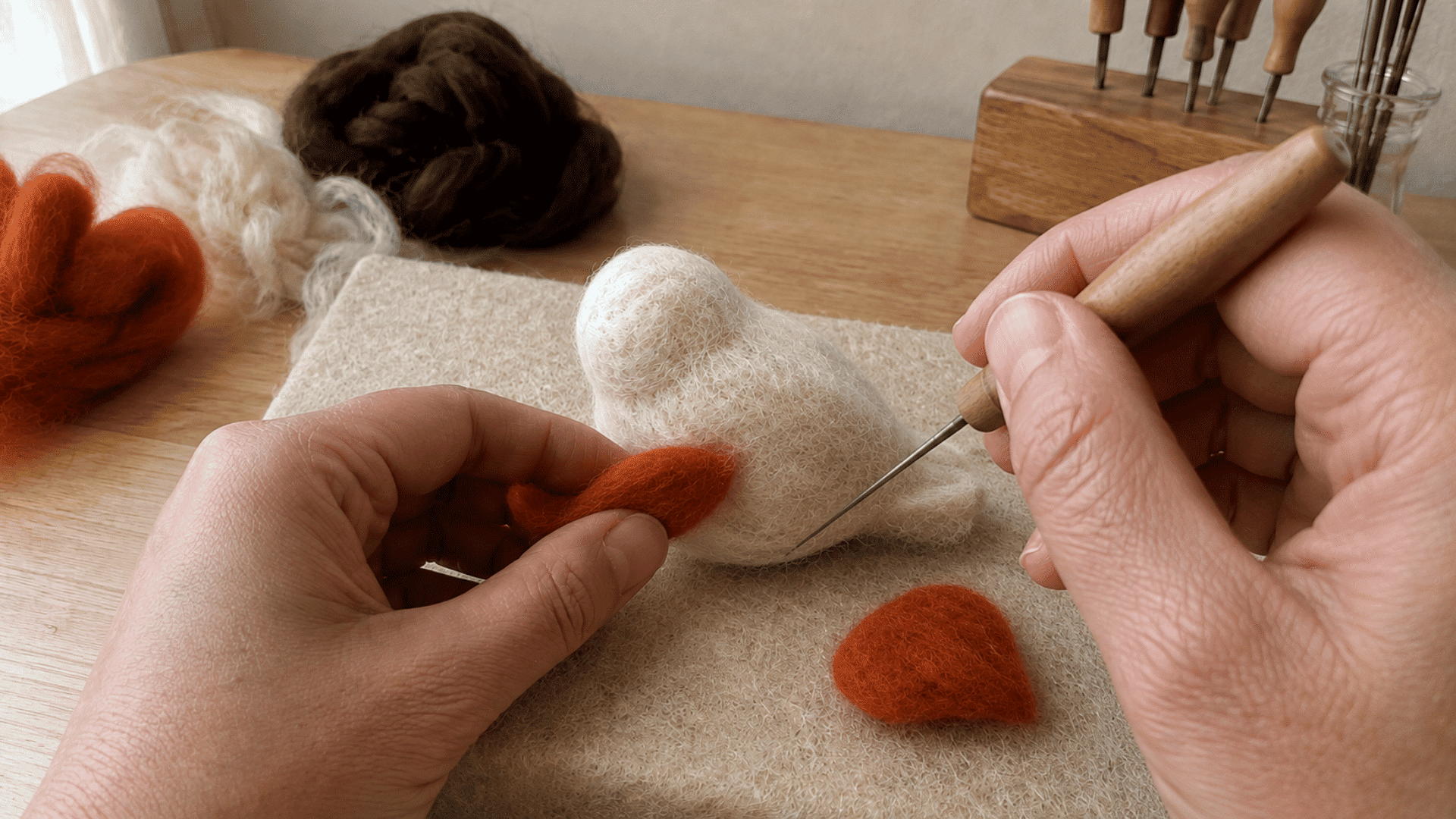 Hands attaching a felted orange wing piece onto a white bird-shaped wool body