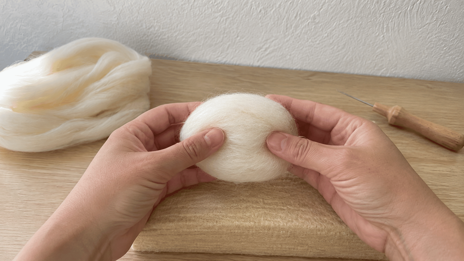 Hands rolling loose white wool into a rounded ball shape on a wooden felting mat