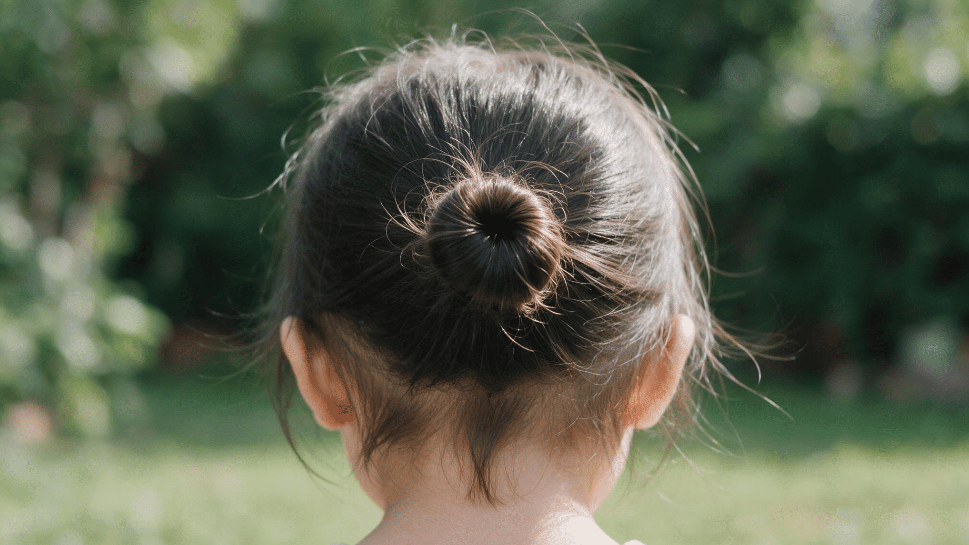 little girl with a sleek ballet bun hairstyle viewed from the back outdoors with a few soft wispy strands around the nape of her neck