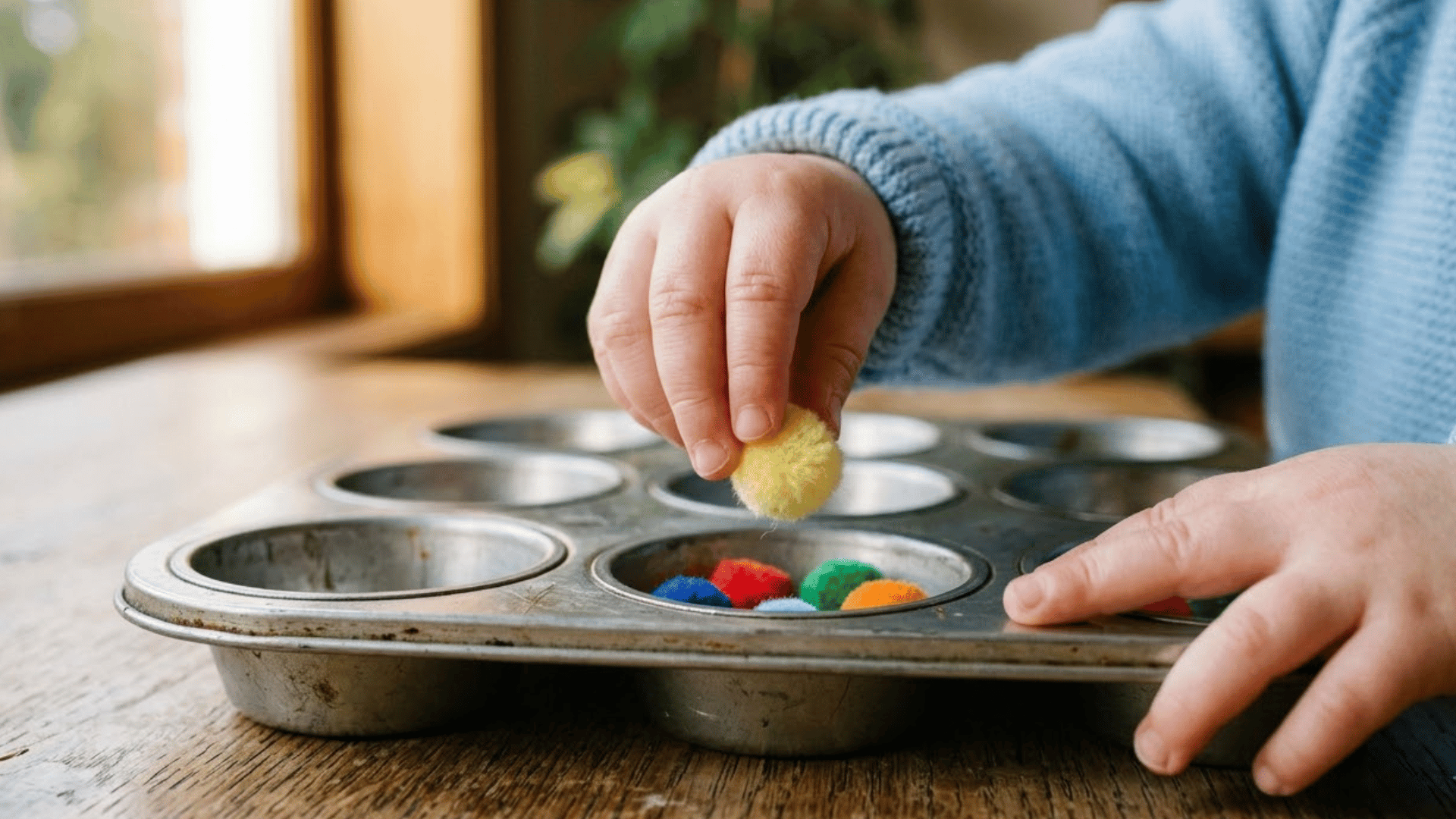 toddler hand pinches a pom pom above a metal muffin tin cup for transfer
