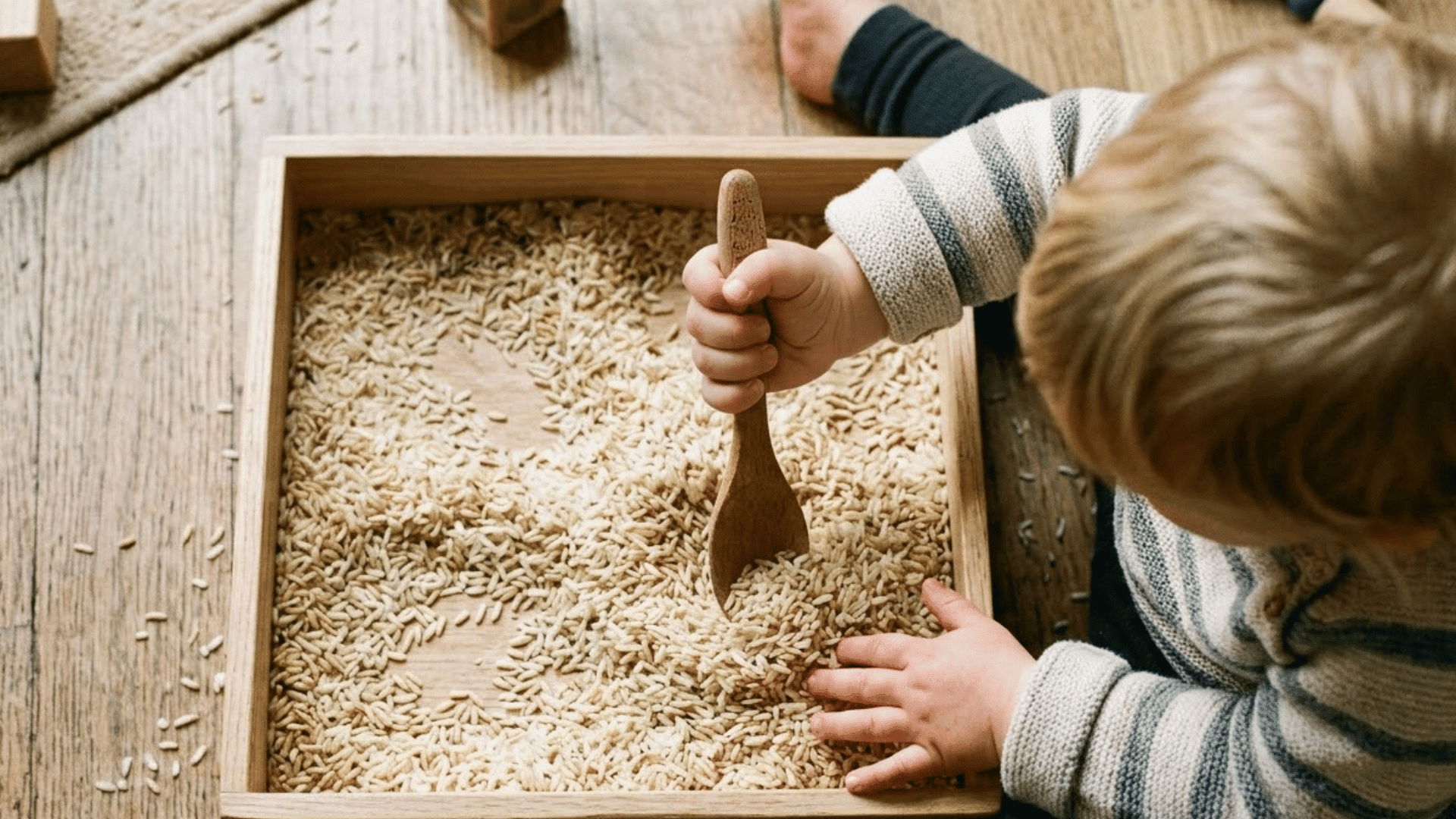 toddler hand grips a wooden spoon while scooping dry rice in a shallow sensory bin