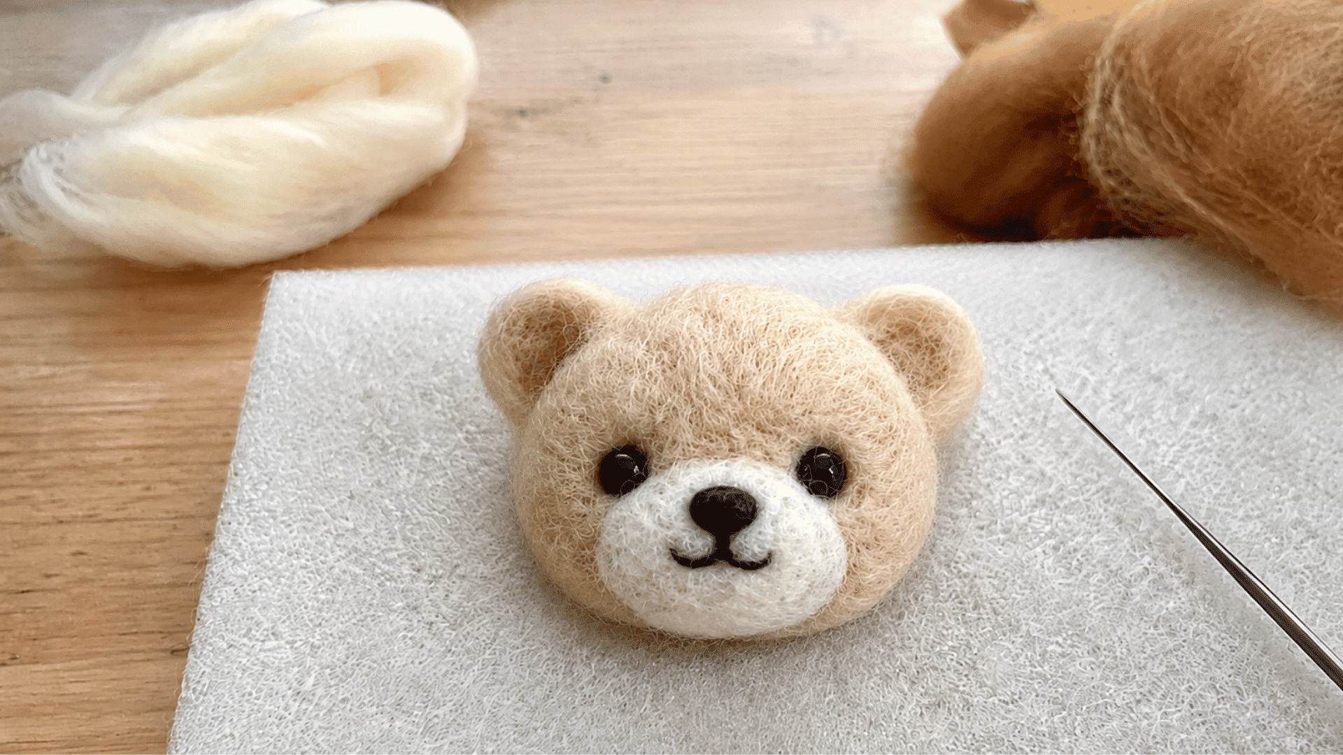 Small felted bear head with ears, nose, and eyes on a felting mat with needle