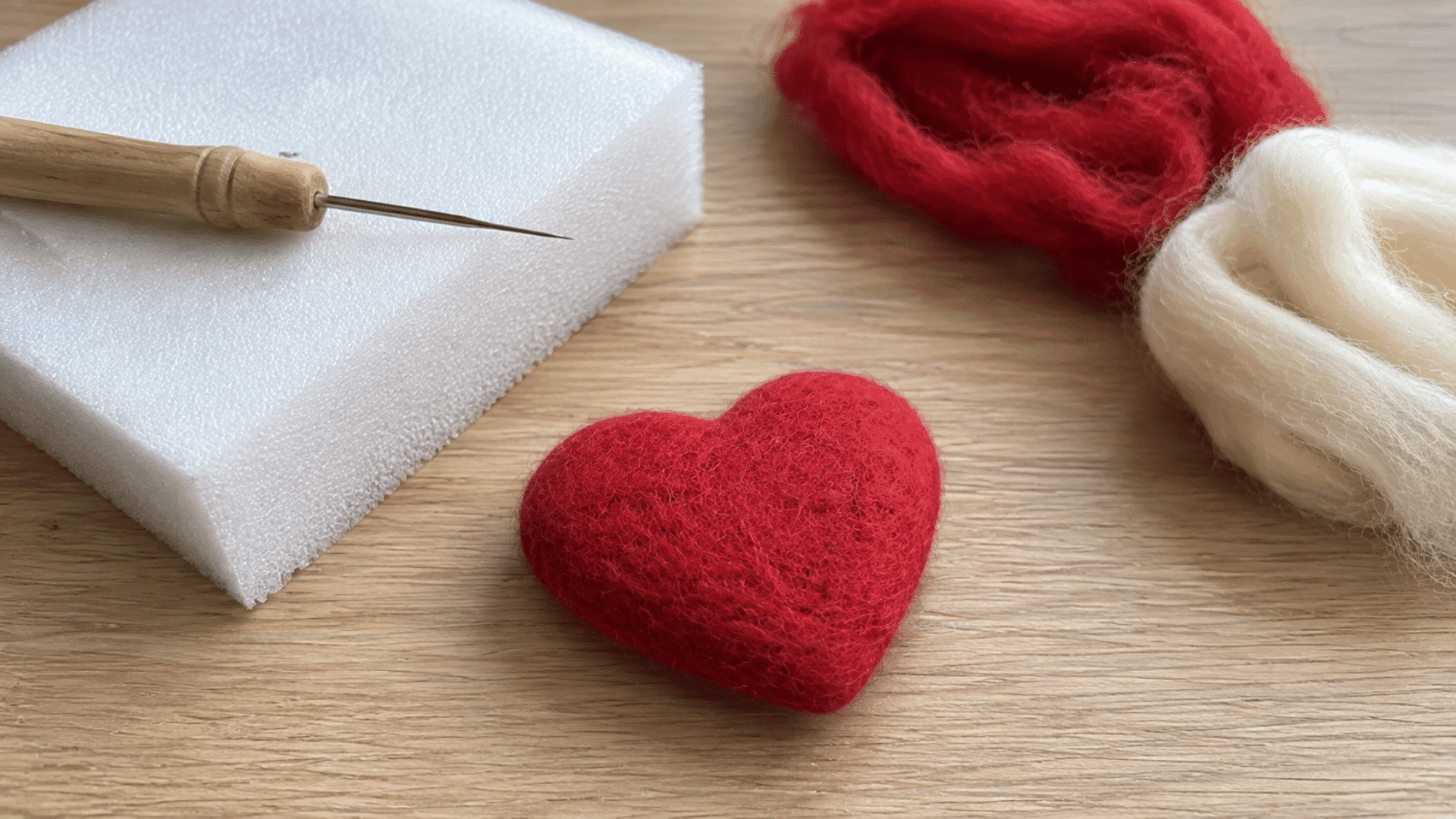 Small felted red heart shape on wooden table with felting needle and wool roving