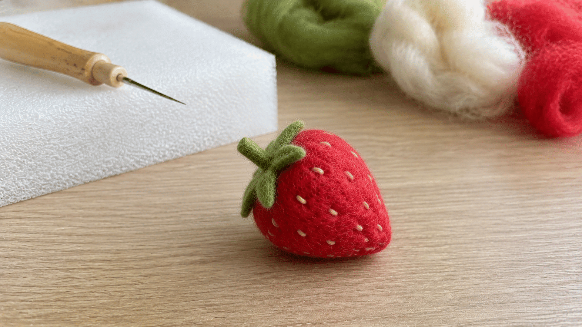 Small felted red strawberry with green leafy top, felting needle nearby