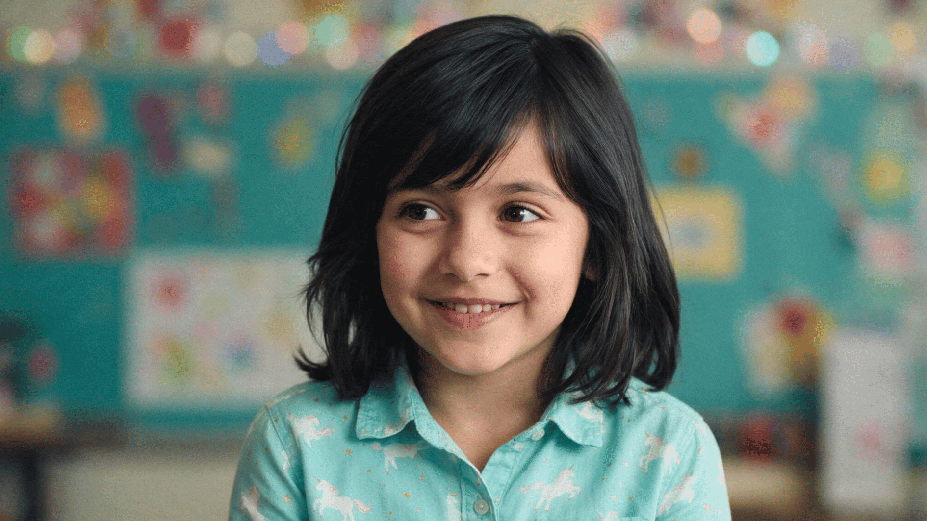 smiling child with shoulder length flipped ends lob hairstyle dark hair sitting indoors with colorful background and wearing a patterned shirt