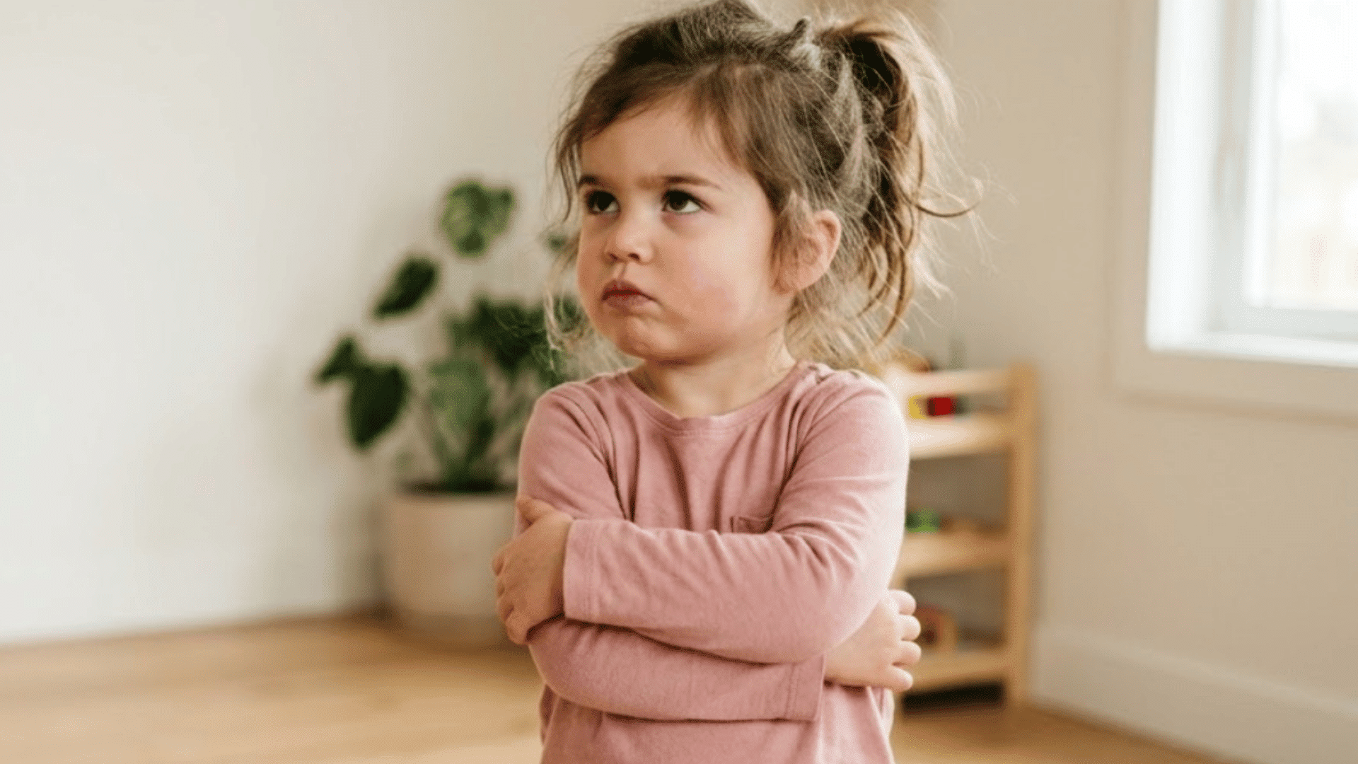 stubborn toddler girl stands with crossed arms in a bright room, looking upward with a serious expression