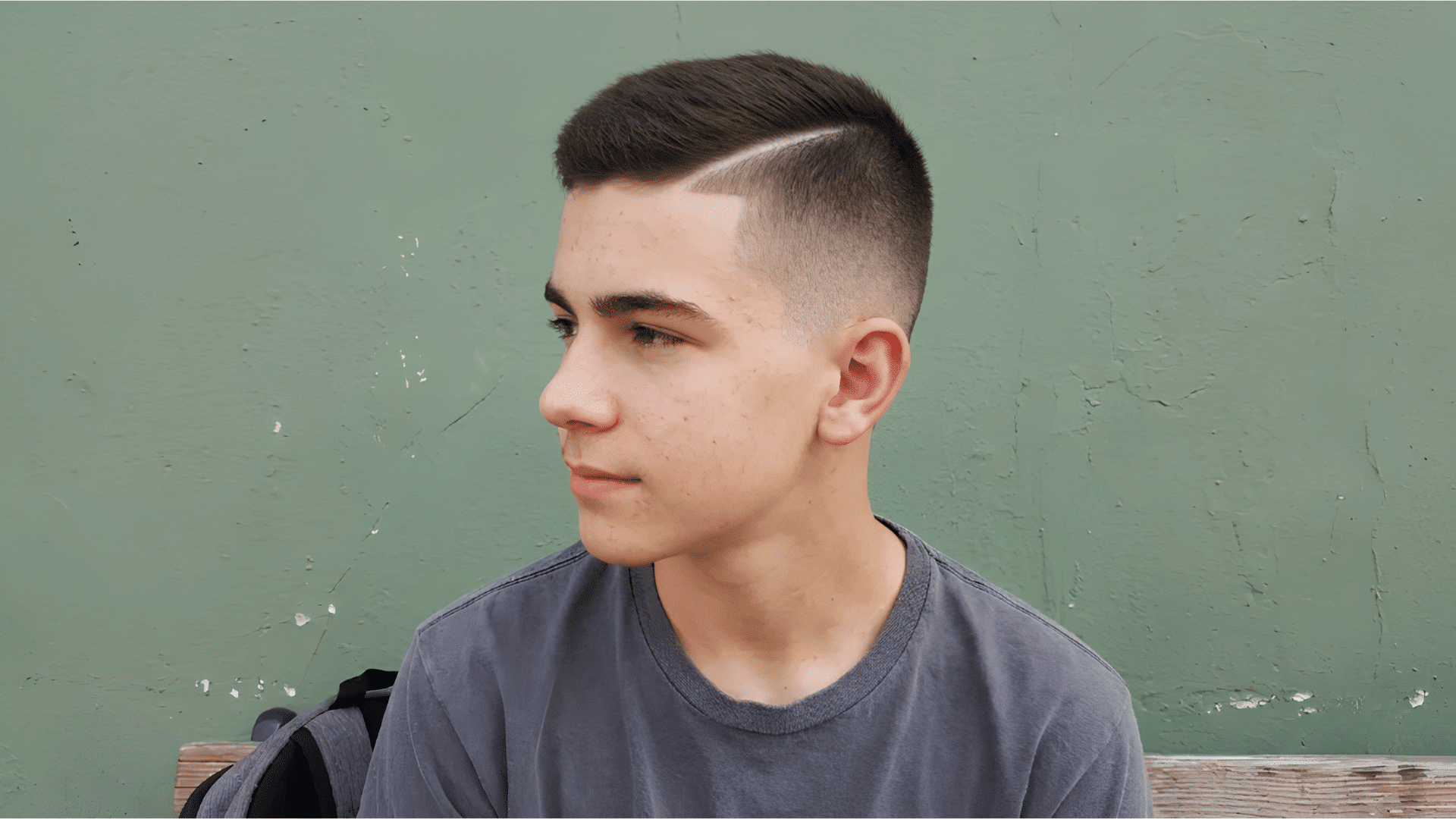 teen boy in profile showing crew cut fade with hard side part line against green wall
