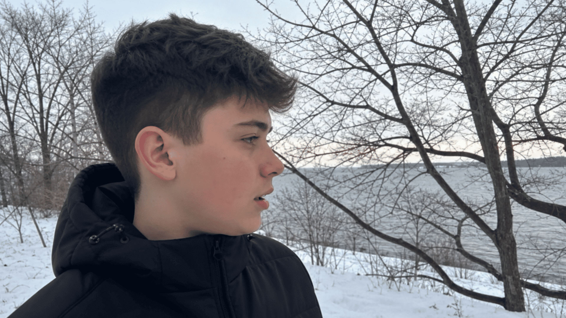 teen boy with winter crew cut longer top for warmth standing near snowy lake shore