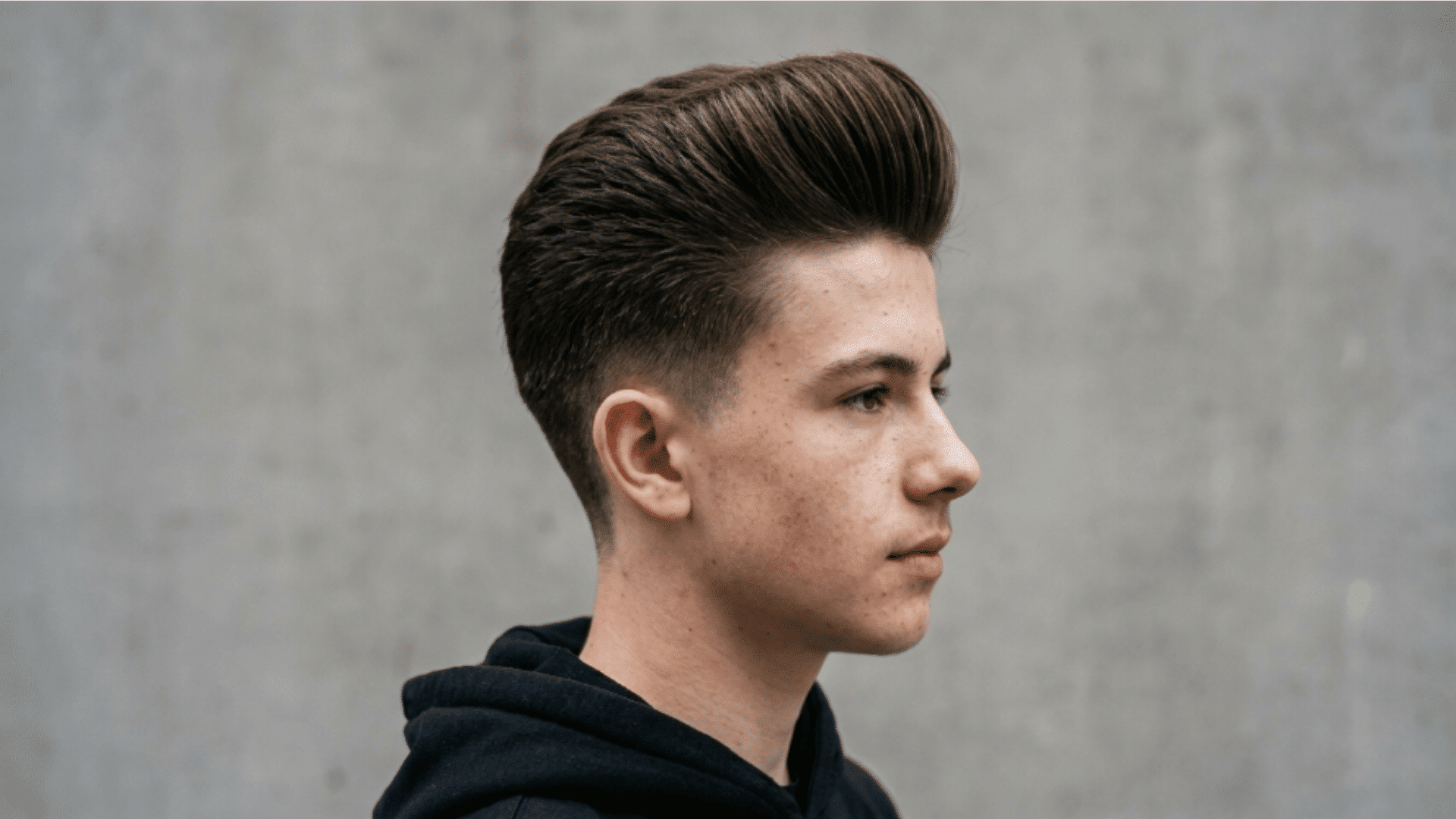 teenage boy in profile with a tall brushed-back pompadour and faded sides, wearing a black hoodie against a grey wall
