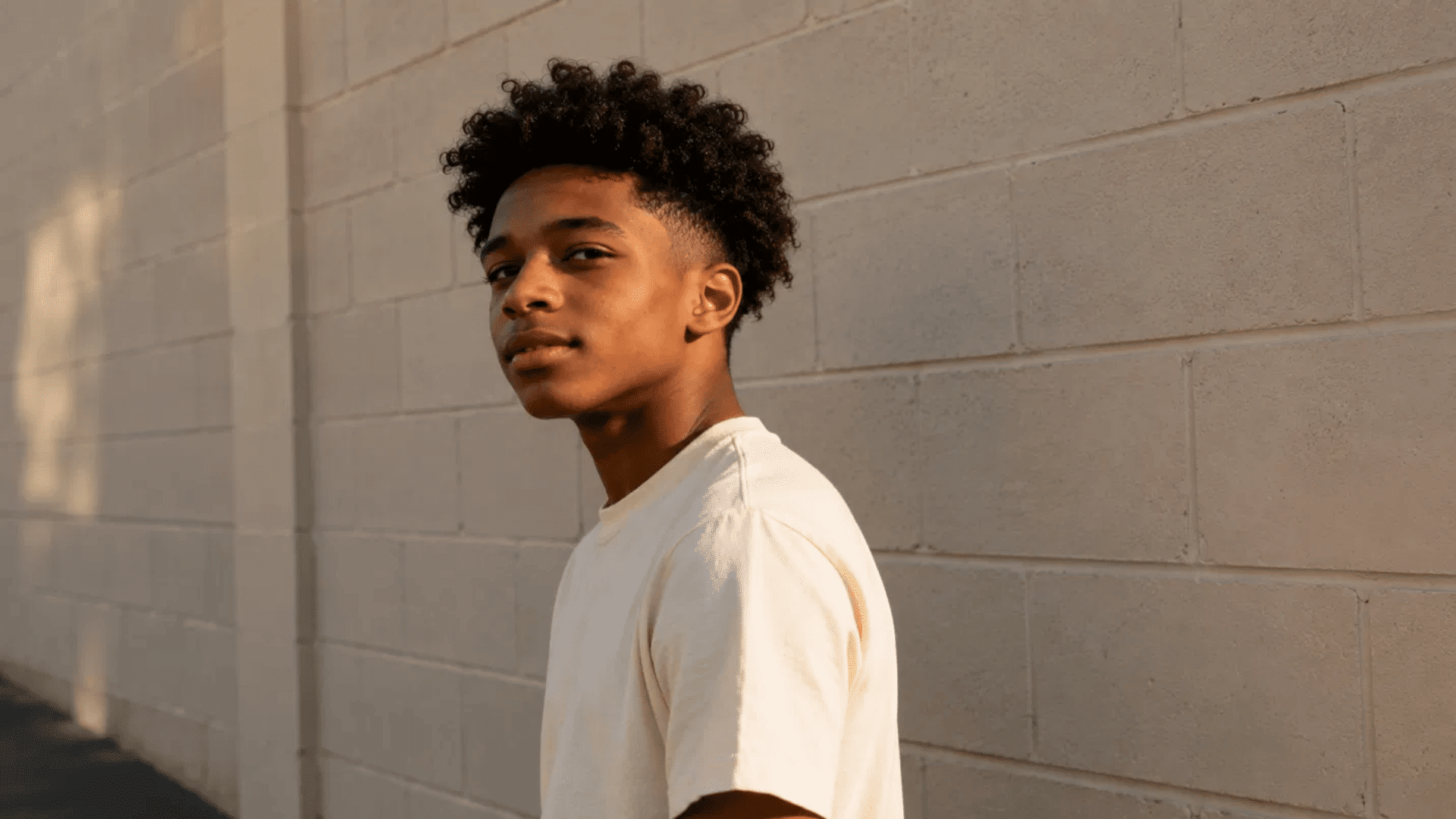 teenage boy with a curly top fade hairstyle wearing a cream t-shirt, standing against a beige brick wall in warm light