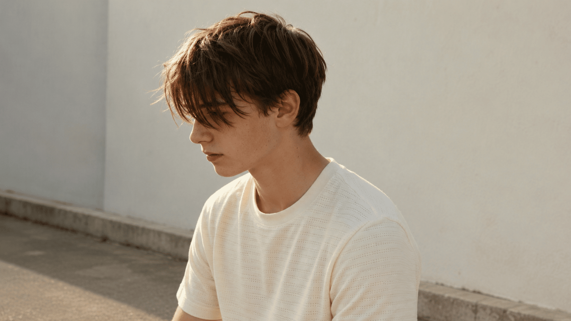 teenage boy with a messy fringe hairstyle looking down, wearing a cream t-shirt against a white wall in warm sunlight