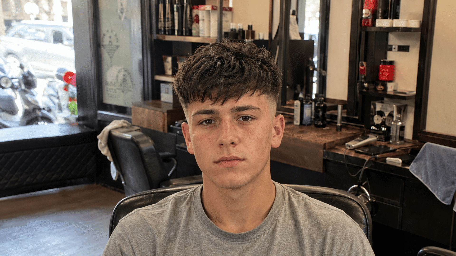 teenage boy with a textured crop fringe and faded sides sitting in a barbershop chair, styling products and mirrors behind him