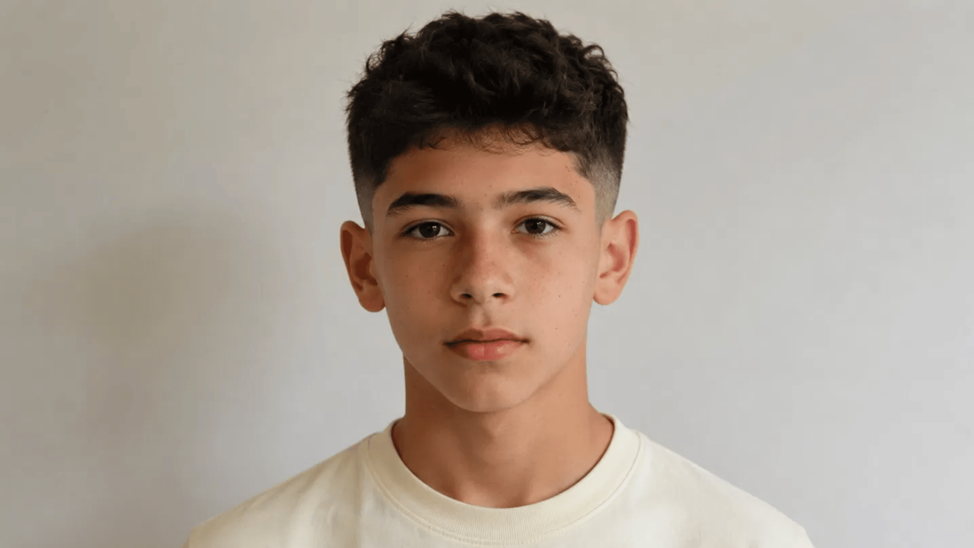 teenage boy with curly low fade haircut facing forward, plain gray background, cream sweatshirt, neutral expression